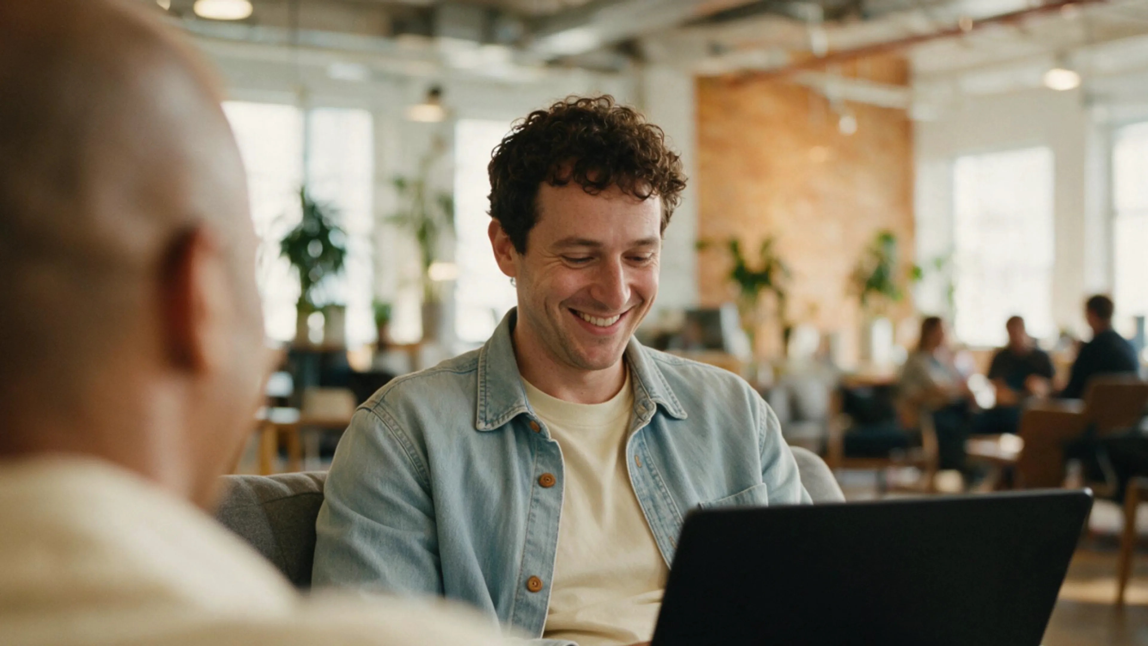 A man with curly hair smiles at his laptop in a bright, modern office. Another person sits nearby, blurred in the foreground. Plants and people are visible in the background.