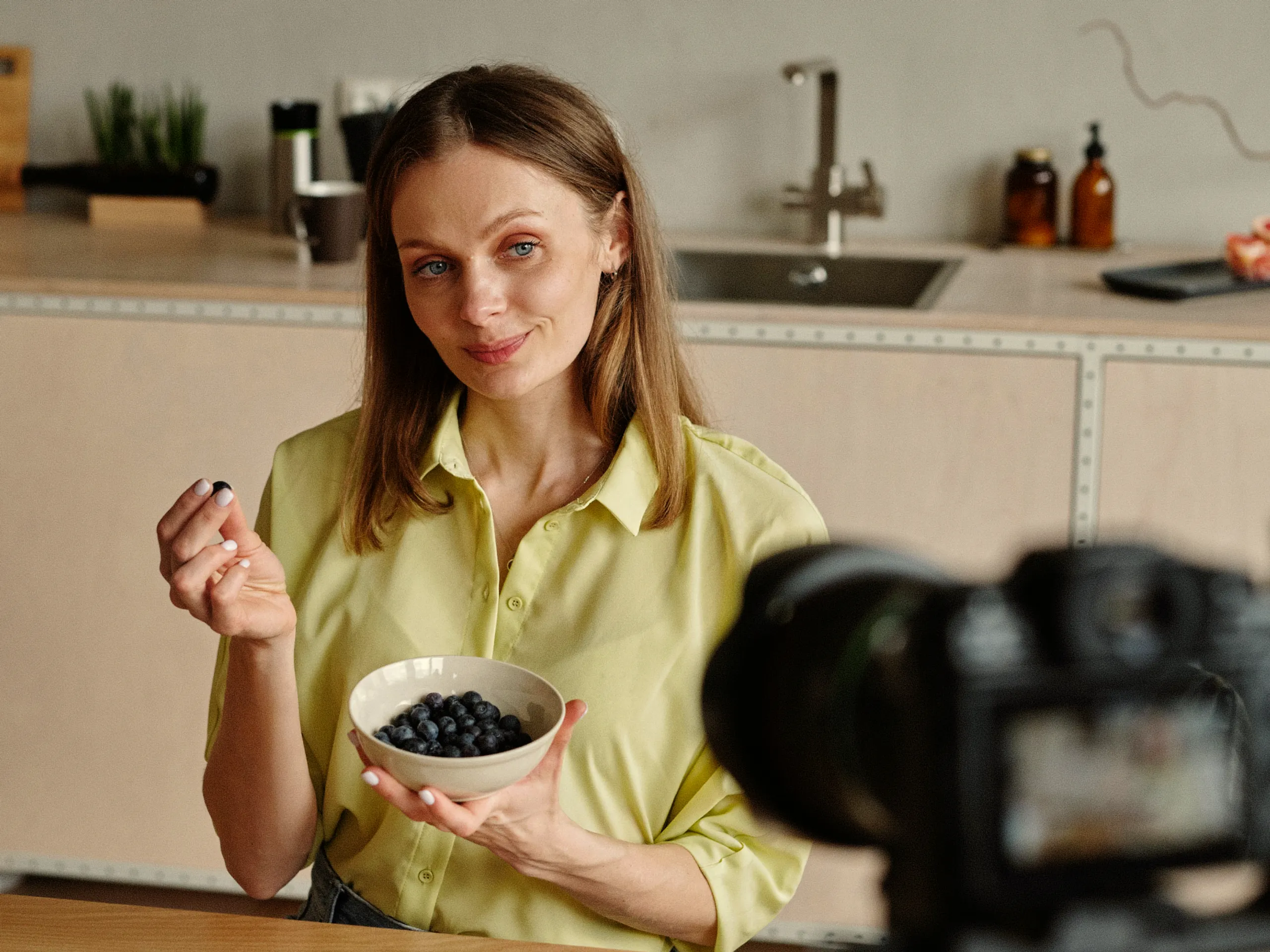 A woman in a yellow shirt holds a bowl of blueberries and a single berry, sitting at a kitchen counter while facing a camera, as if filming or taking a photo.