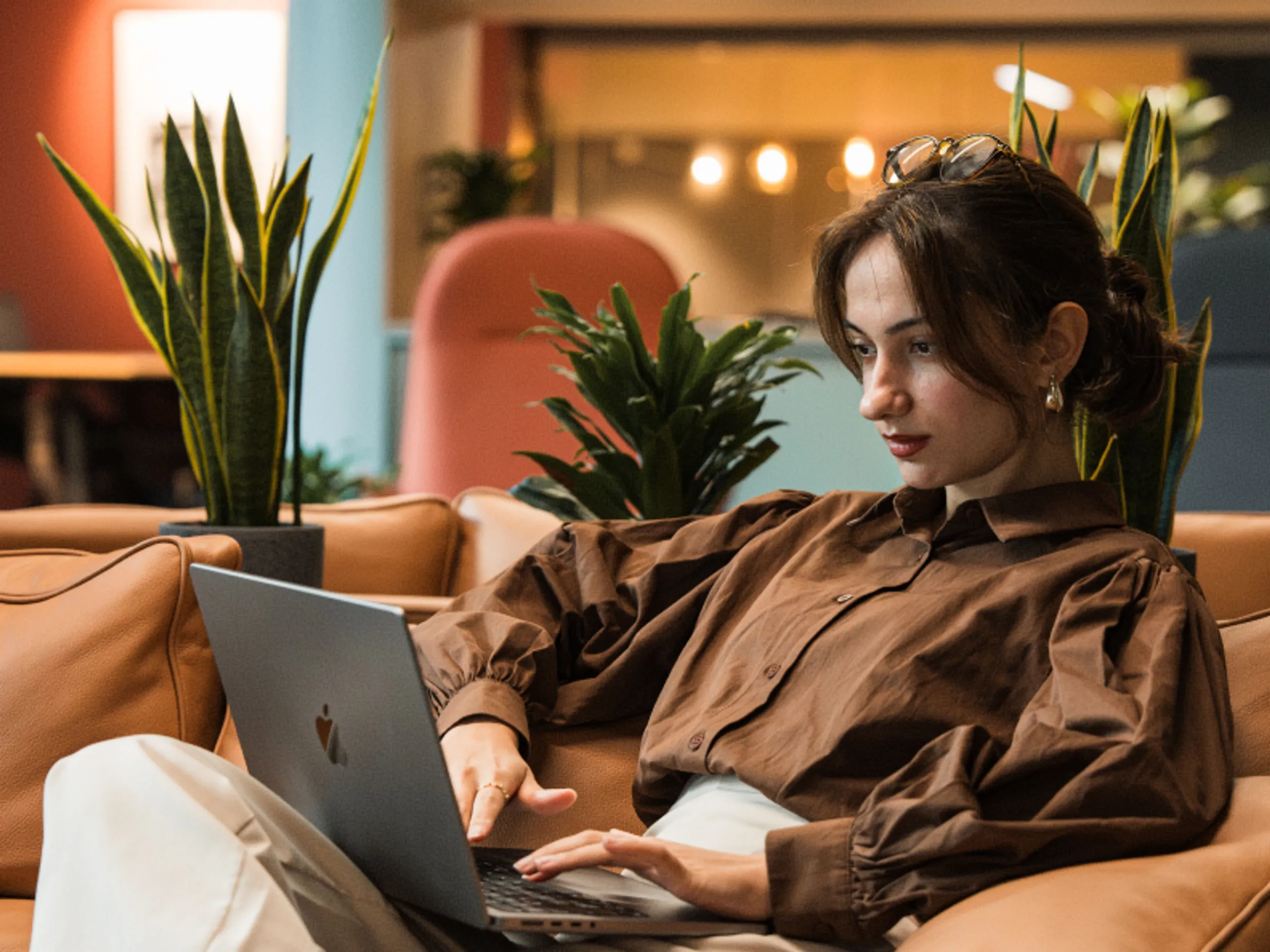 A woman with brown hair, wearing a brown blouse, sits on a tan couch using a laptop. She appears focused, surrounded by green plants in a modern, warmly lit space.