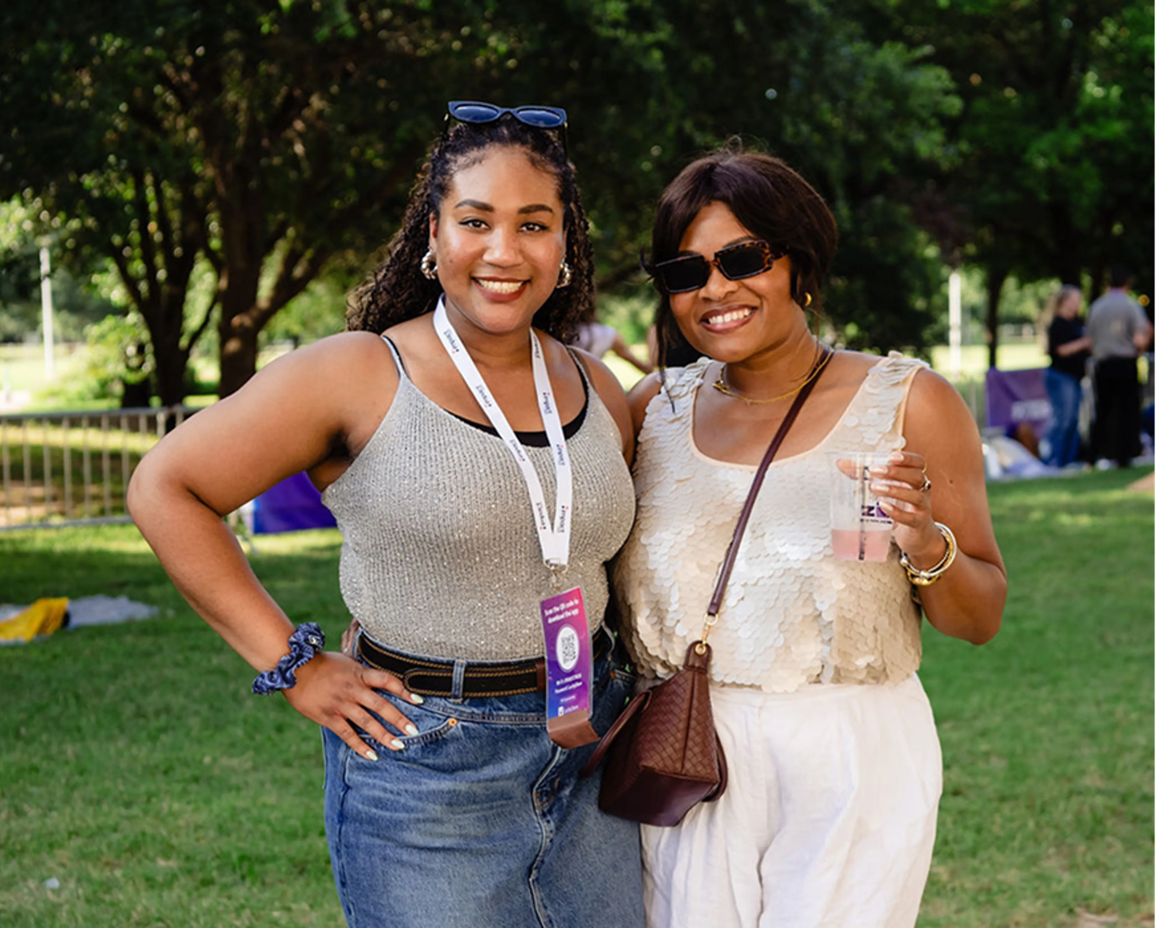 Two women smiling outdoors, wearing summer outfits and sunglasses. One has a lanyard and event badge, the other holds a drink. Green grass and trees are in the background.
