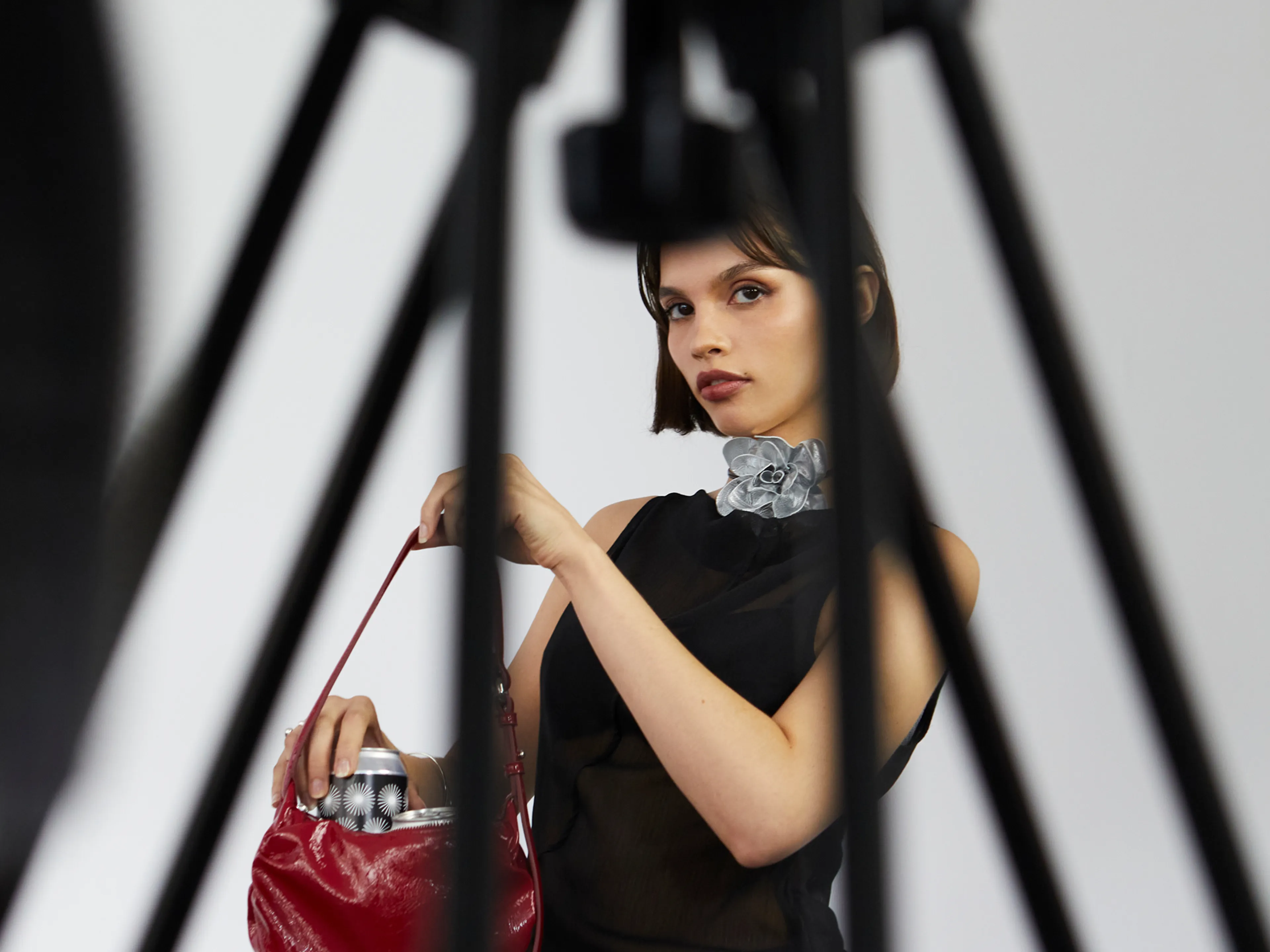 A woman in a black sleeveless top holds a red handbag, looking at the camera through the legs of a tripod in the foreground.