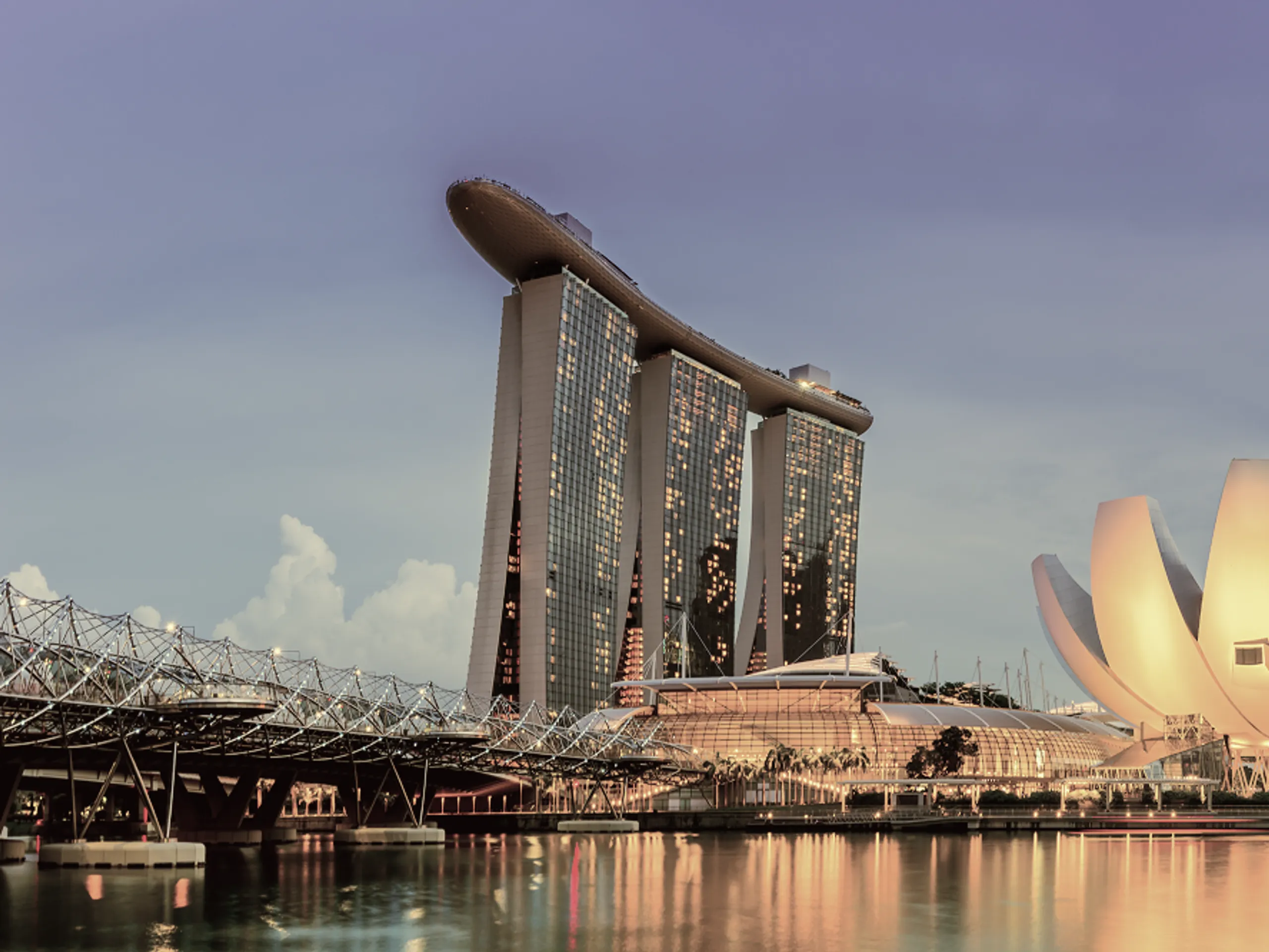 Marina Bay Sands hotel with its three towers and boat-shaped rooftop is seen at dusk beside water, with the Helix Bridge and the ArtScience Museum in Singapore. The sky is light purple and reflects on the calm water.
