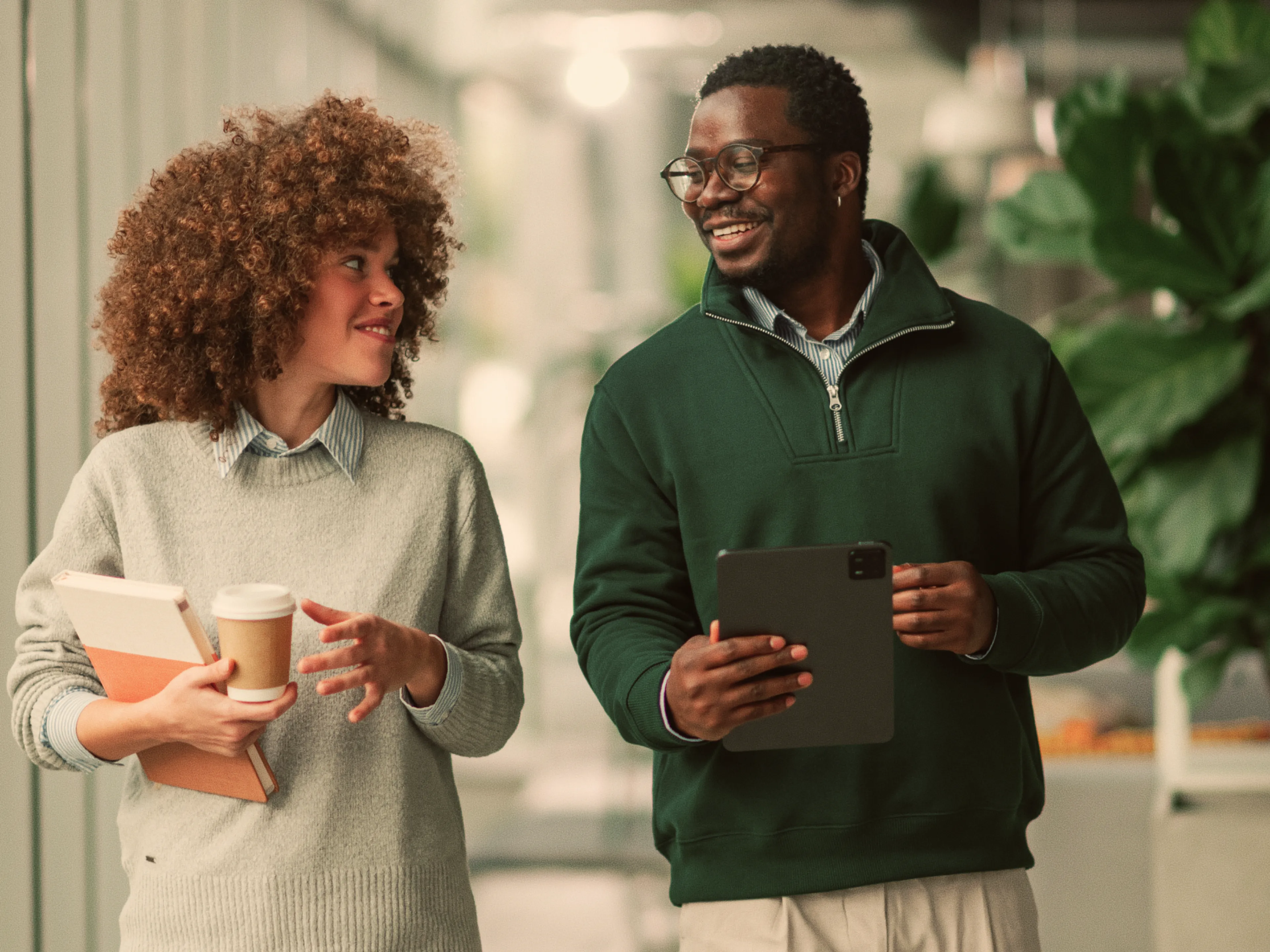 Two colleagues walk indoors, smiling at each other. One holds a coffee cup and notebook, the other holds a tablet. Both wear casual business attire. Large green plants are visible in the background.