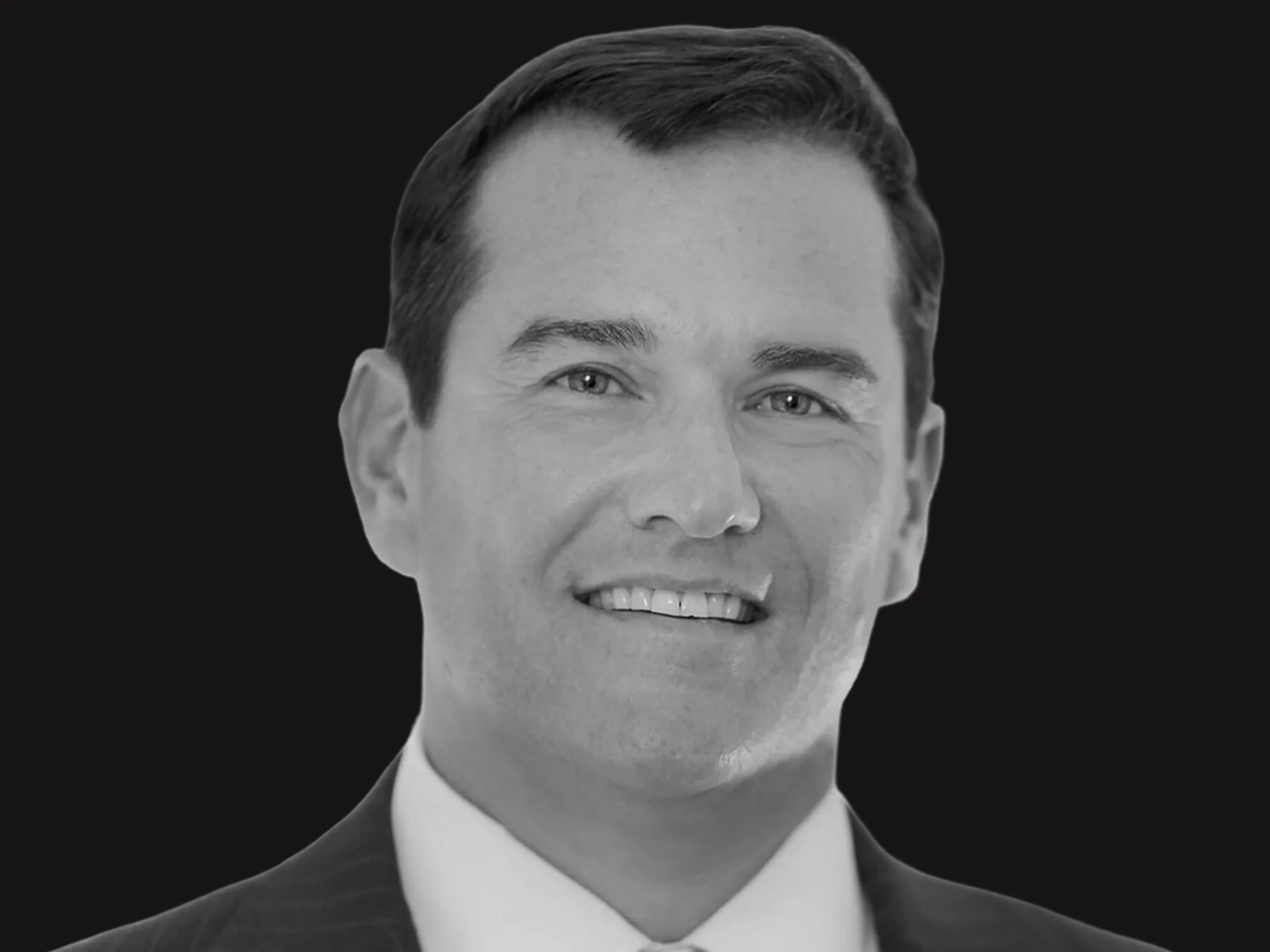 Smiling man in a suit and tie, with short dark hair, seen in a black and white portrait against a solid black background.