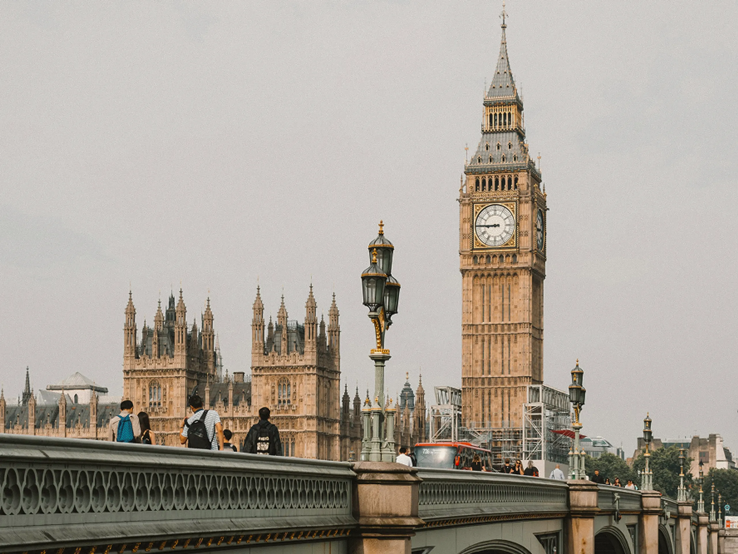 People walk across Westminster Bridge in London. The Houses of Parliament and the Elizabeth Tower, commonly known as Big Ben, are visible in the background on a cloudy day.