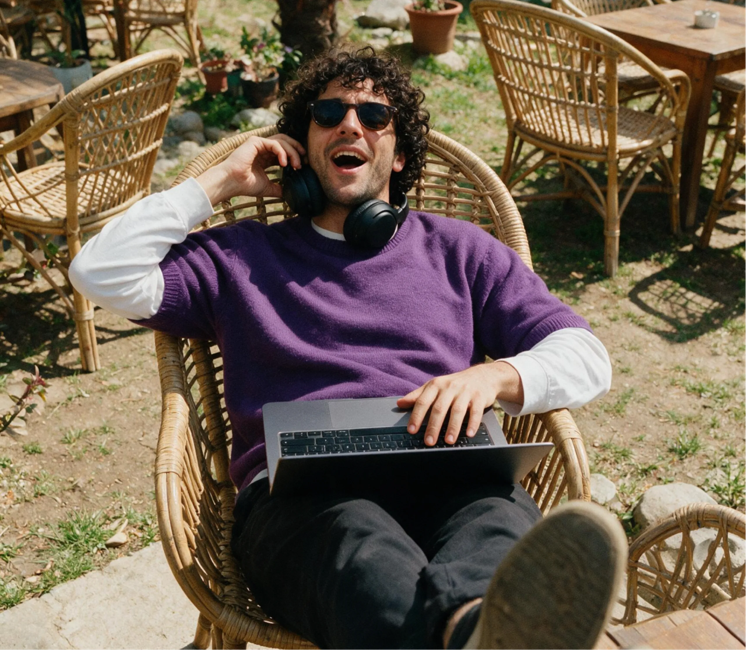 A person wearing sunglasses and headphones relaxes in a wicker chair outdoors, smiling while using a laptop on their lap. There are empty tables and plants in the background.