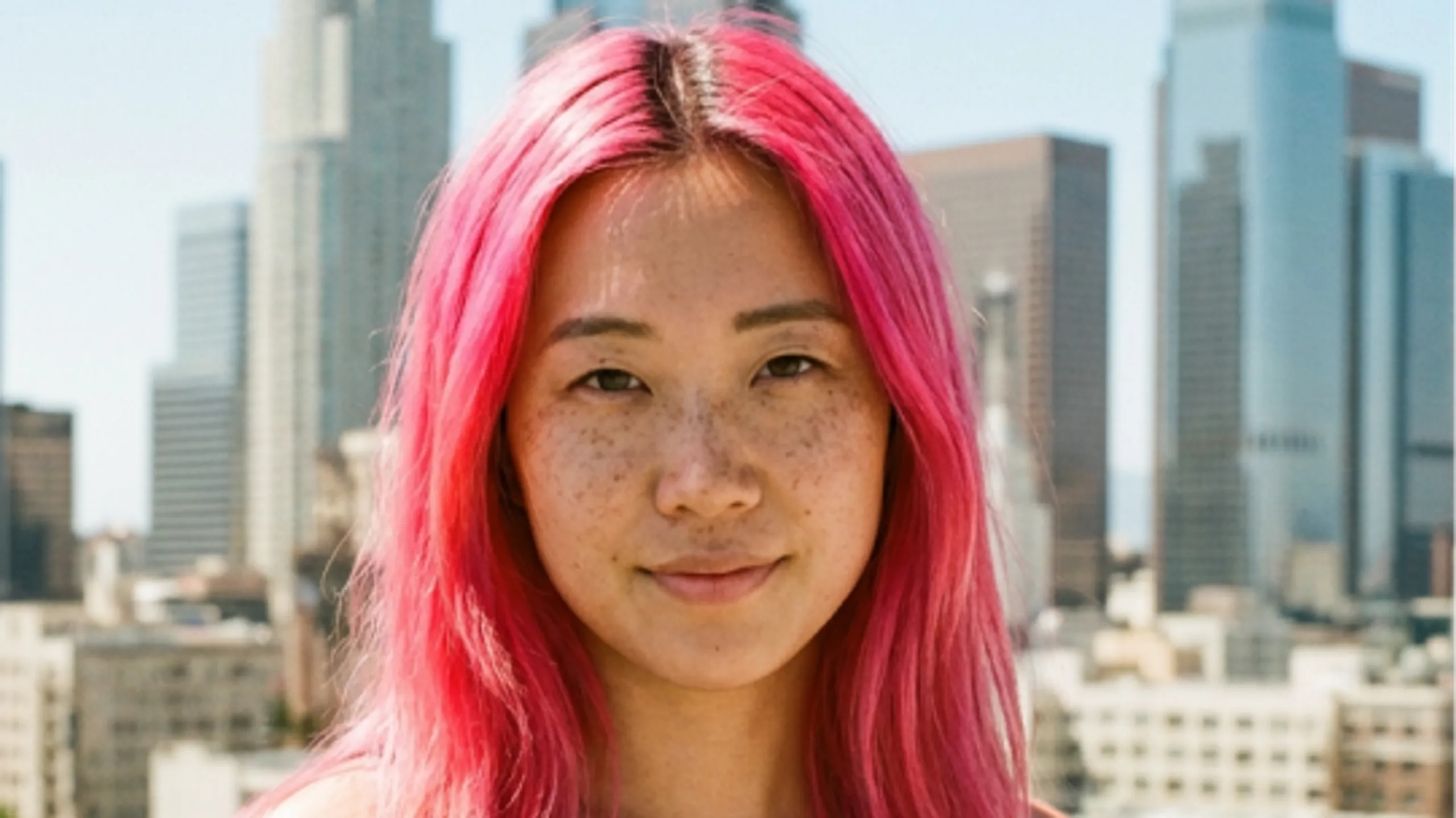 A person with bright pink hair and freckles stands outside with a city skyline and tall buildings in the background on a sunny day.