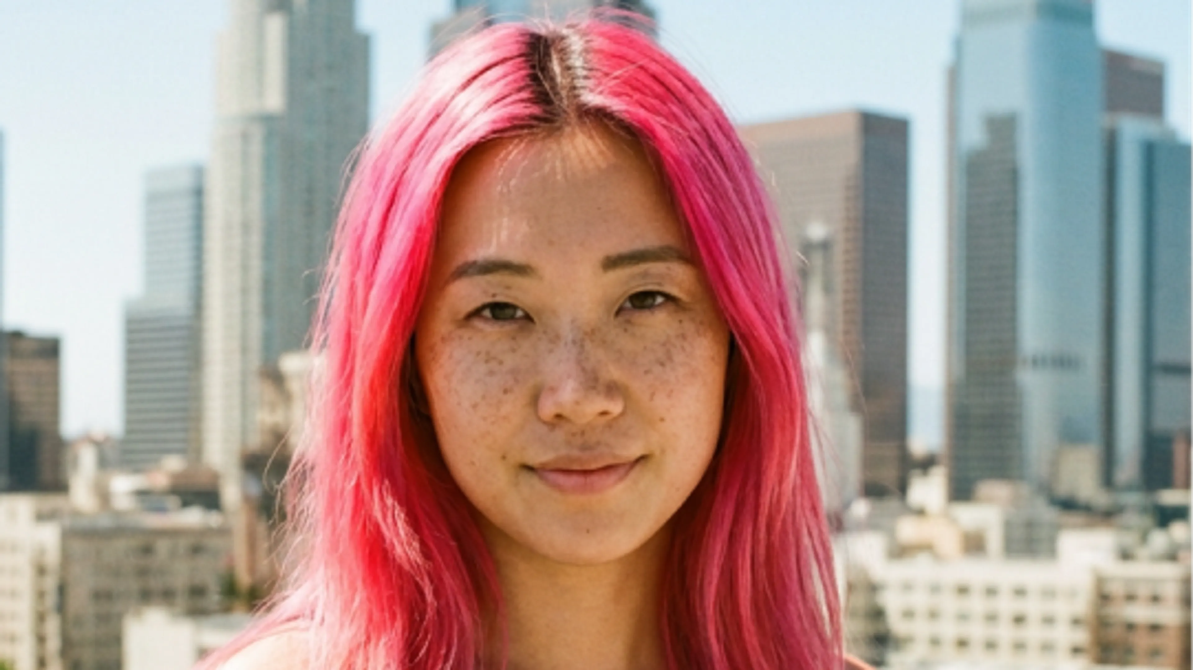 A person with bright pink hair and freckles stands outside with a city skyline and tall buildings in the background on a sunny day.
