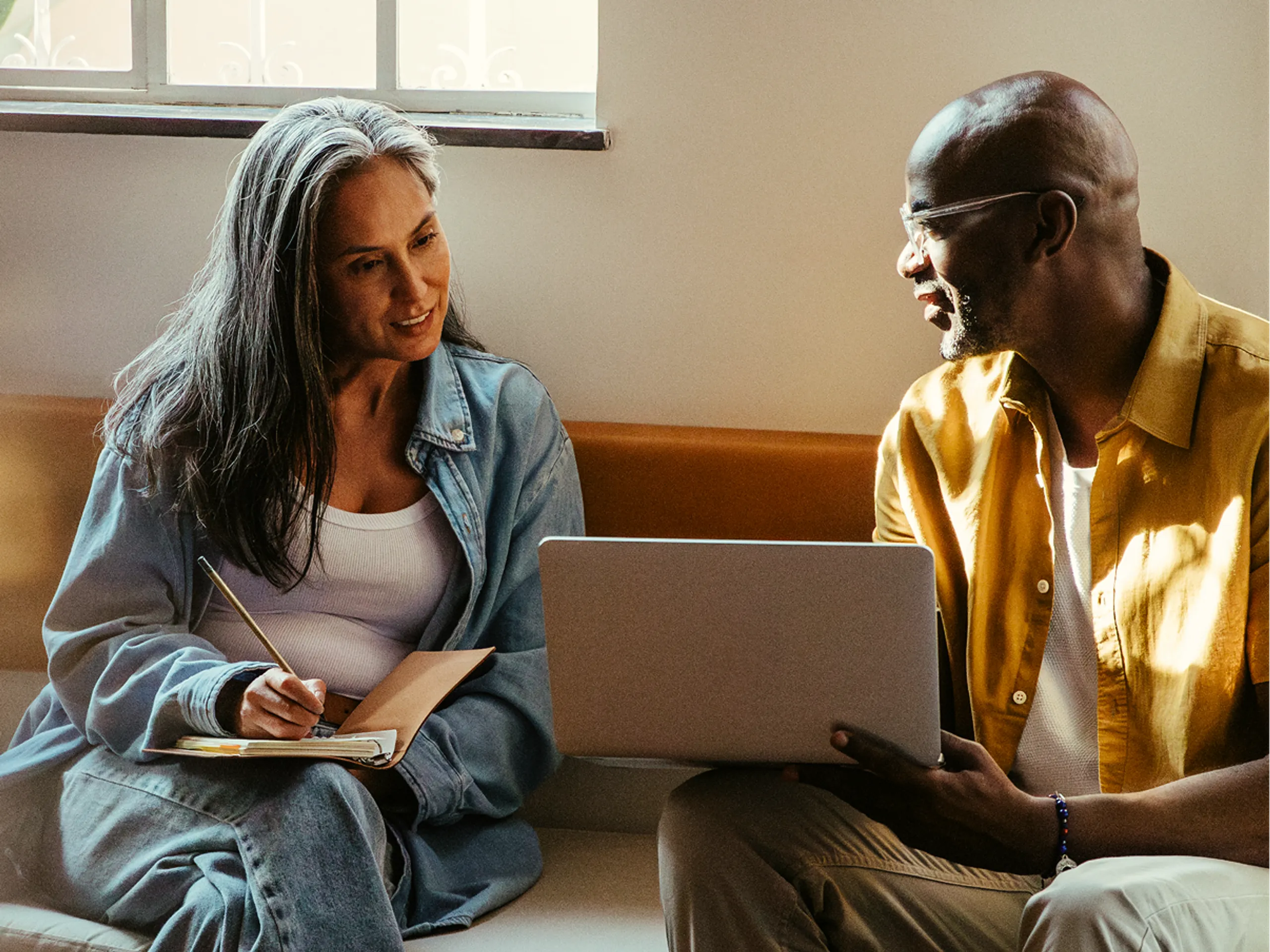 Two people sit on a couch having a discussion. One holds a notebook and pen, while the other holds a laptop. Sunlight streams through a nearby window, creating a warm and collaborative atmosphere.