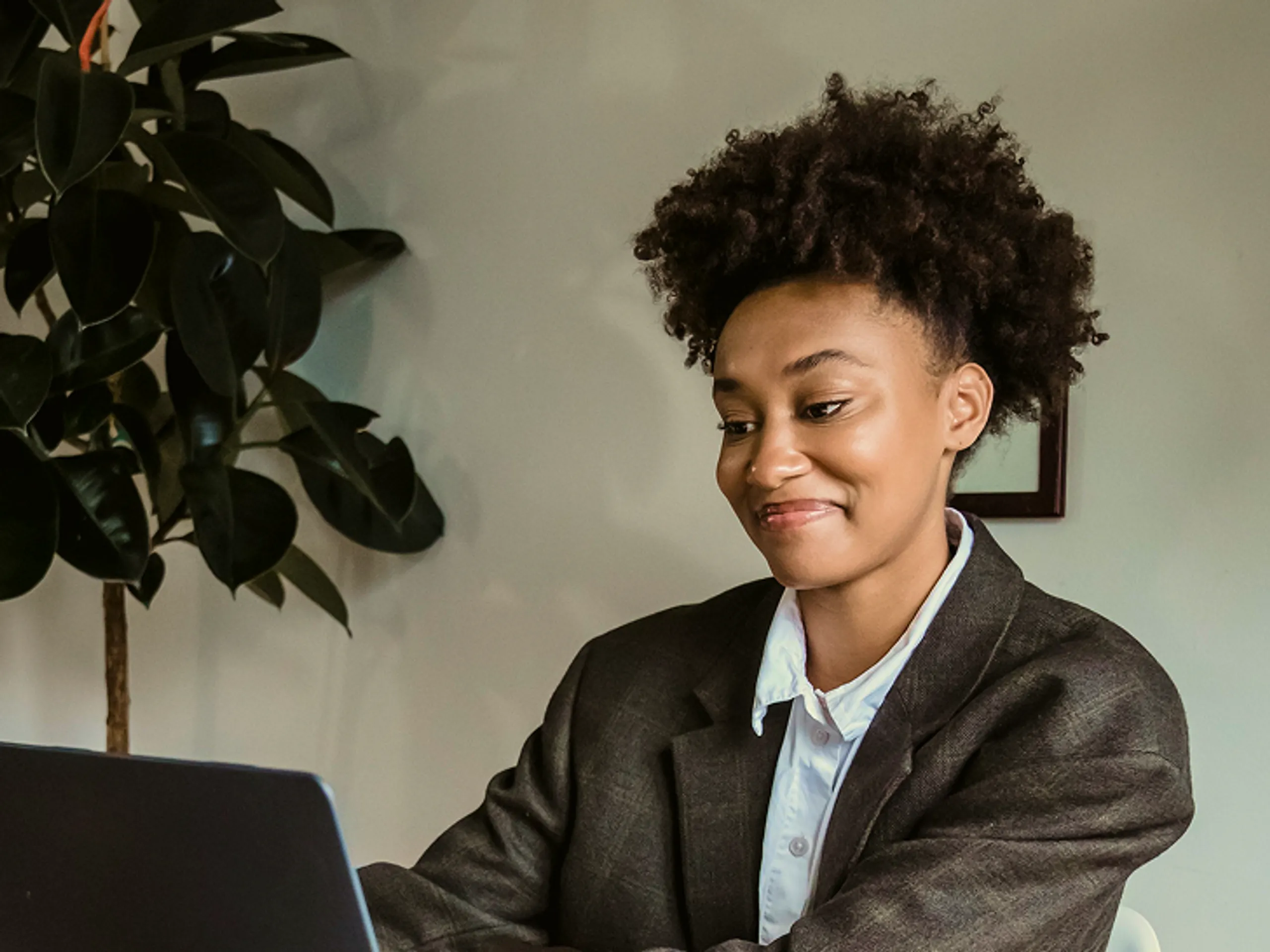 A person with curly hair wearing a suit jacket and white shirt sits at a desk, smiling while looking at a laptop. A leafy indoor plant is visible in the background.