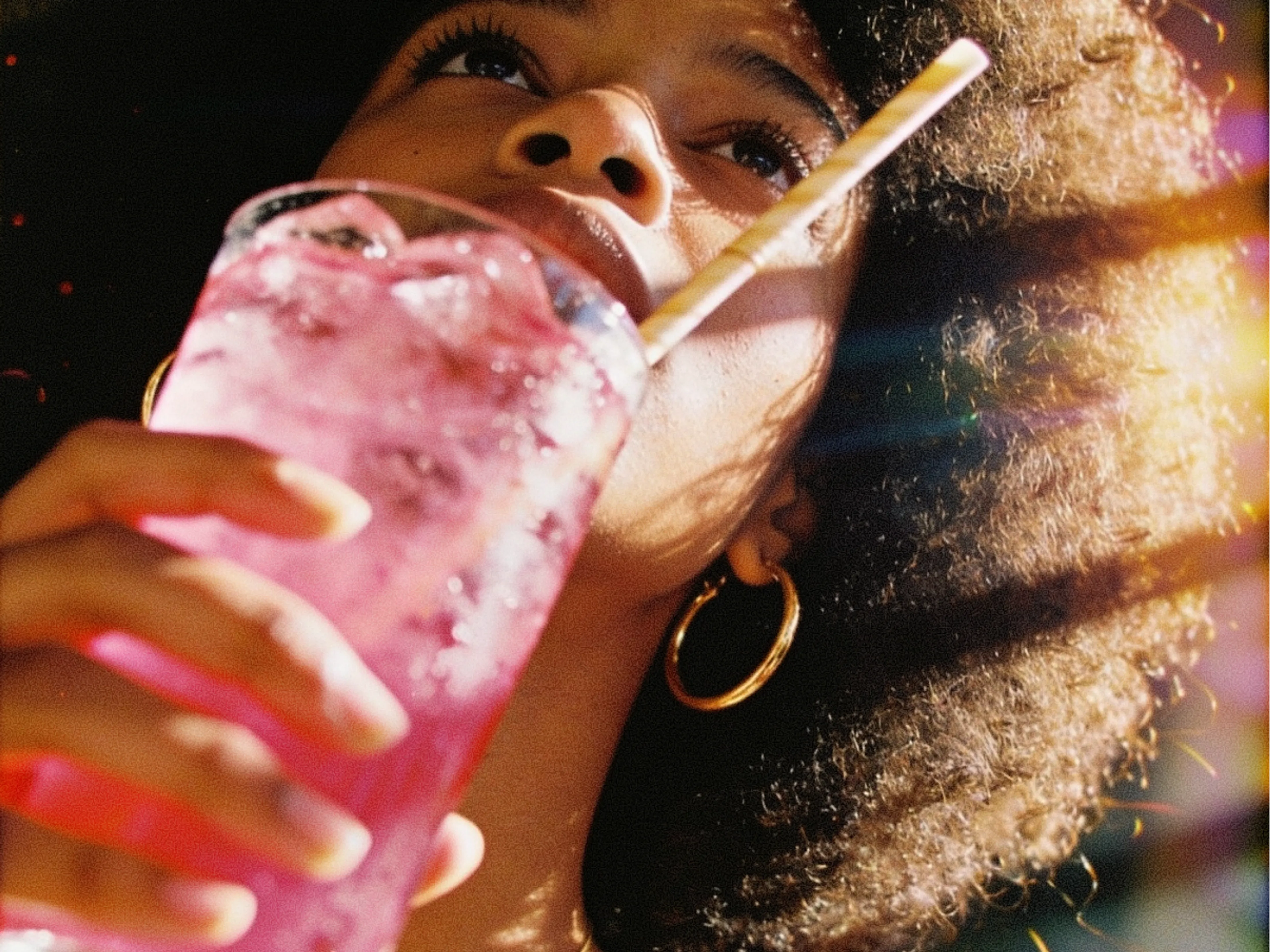 A person with curly hair and gold hoop earrings drinks a cold, pink beverage with ice through a straw, sunlight highlighting their face.