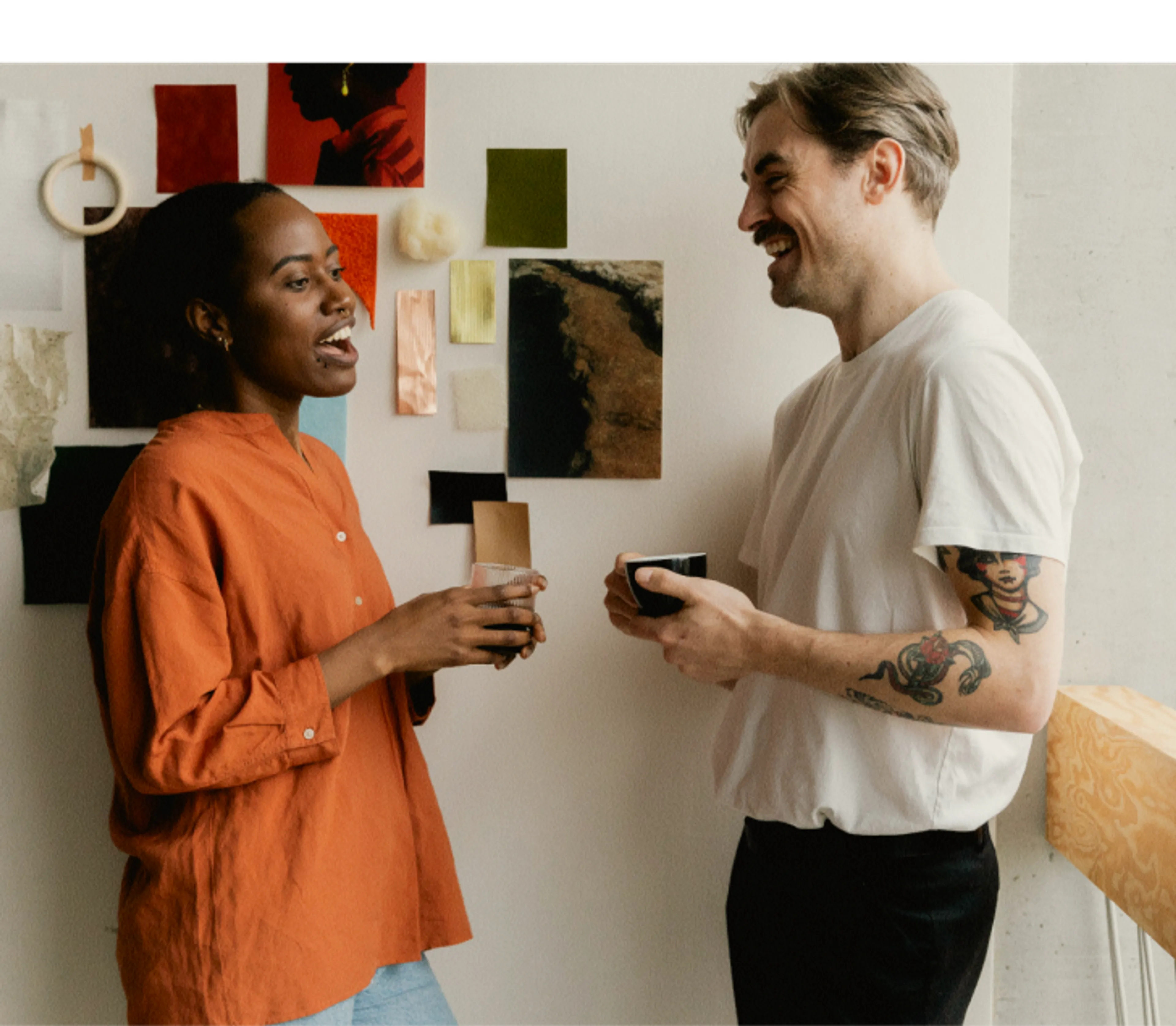 Two people stand indoors, smiling and talking while holding cups. Behind them is a wall decorated with colorful samples and photos. One person wears an orange shirt; the other wears a white shirt and has a visible arm tattoo.