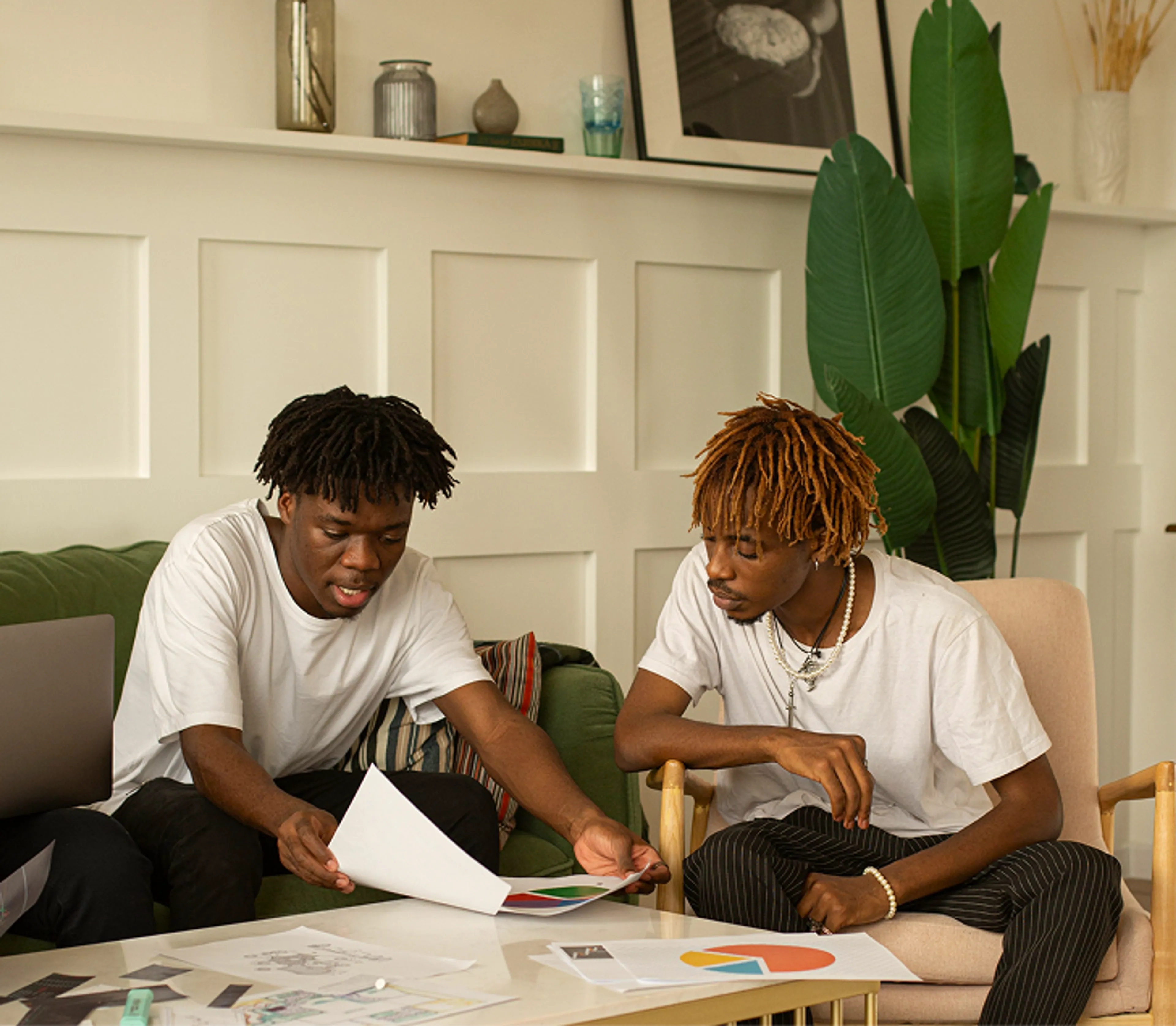 Two young men sit at a coffee table reviewing papers and charts together in a modern, plant-filled room with framed art on the mantel.