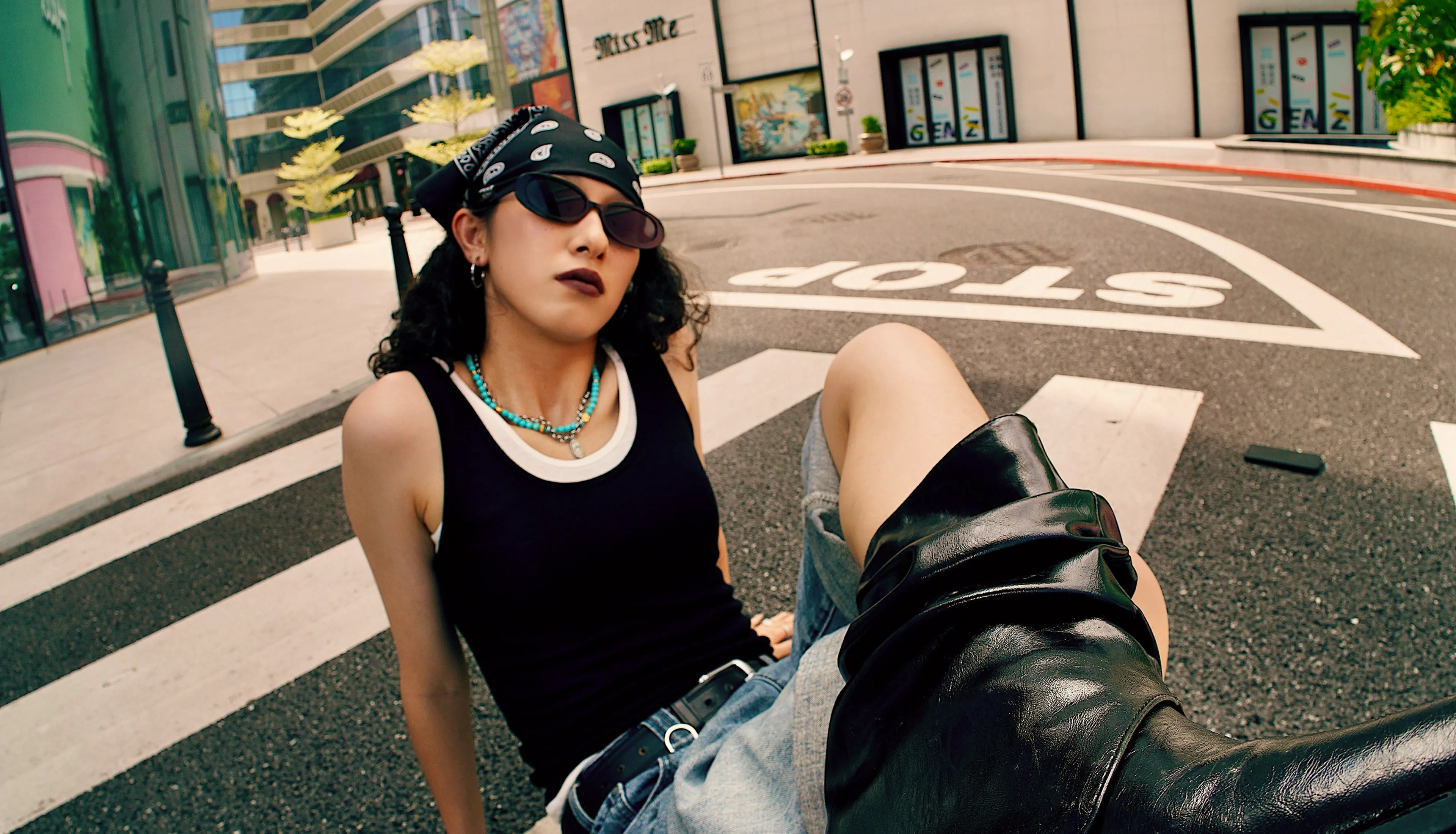 Person wearing a black tank top, bandana, sunglasses, and boots poses confidently on a city street crosswalk near a STOP sign, with buildings and storefronts in the background.