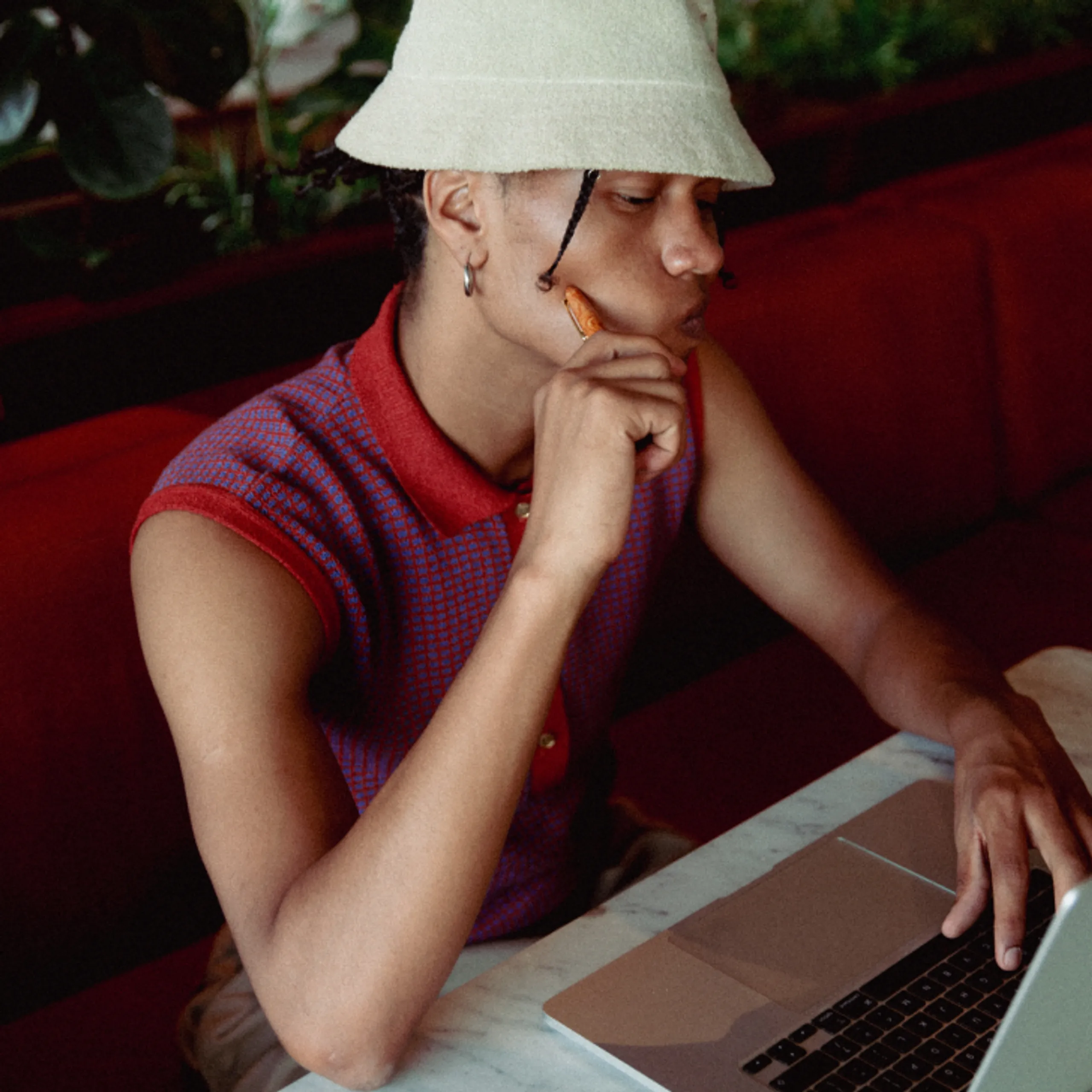 A person wearing a sleeveless red shirt and light bucket hat sits at a table, resting their chin on their hand while looking at a laptop screen, appearing deep in thought.