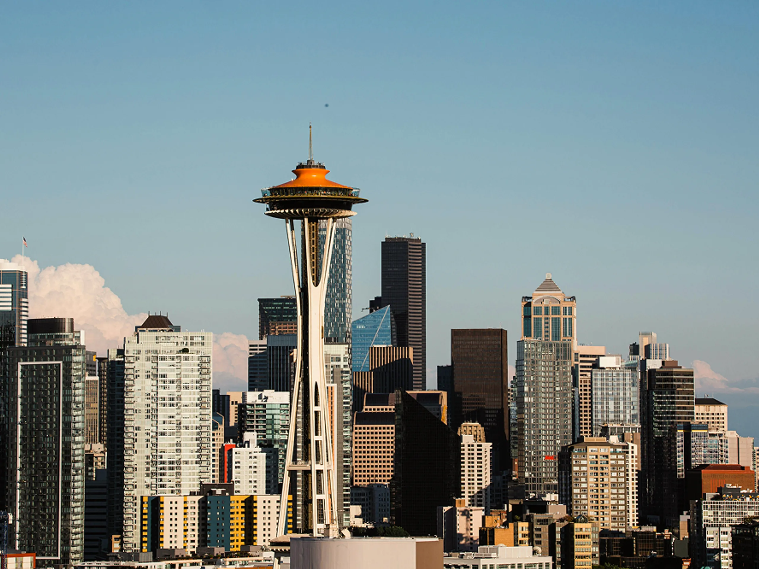 The Space Needle stands in front of the Seattle skyline with modern skyscrapers and a clear blue sky in the background.