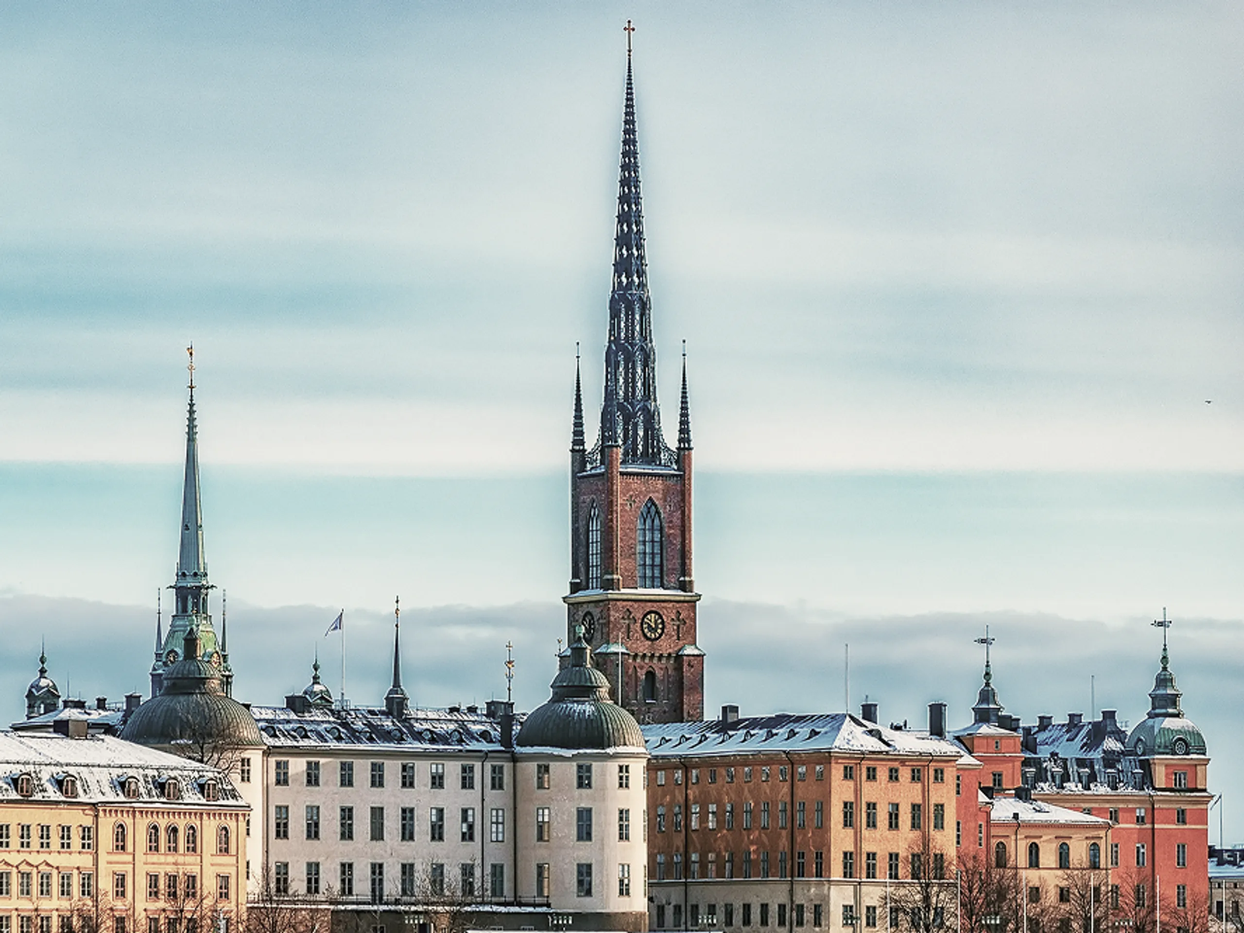 Snow-covered historic buildings with domed roofs stand in front of a tall church with a pointed spire, under a blue sky with thin clouds.