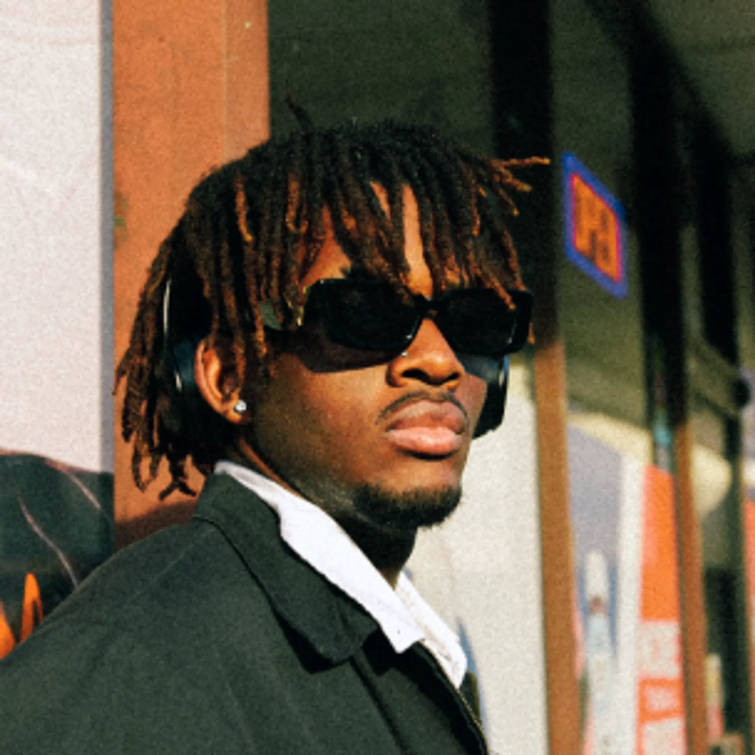 A person wearing sunglasses and headphones stands outside near a glass door with a blue and orange sign in the background. They have short, brown dreadlocks and are dressed in a dark jacket and white collared shirt.