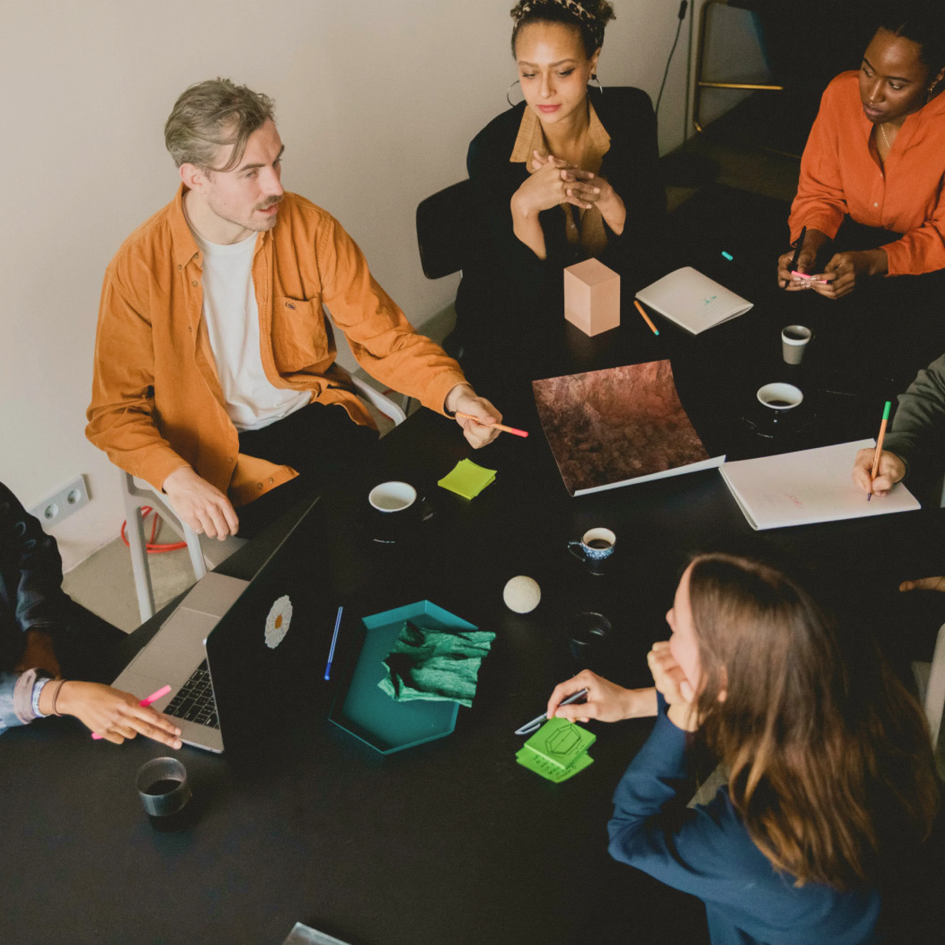 Five people sit around a black table with notebooks, laptops, drinks, and papers, engaged in a discussion. One person gestures while others listen or take notes in a brightly lit room.