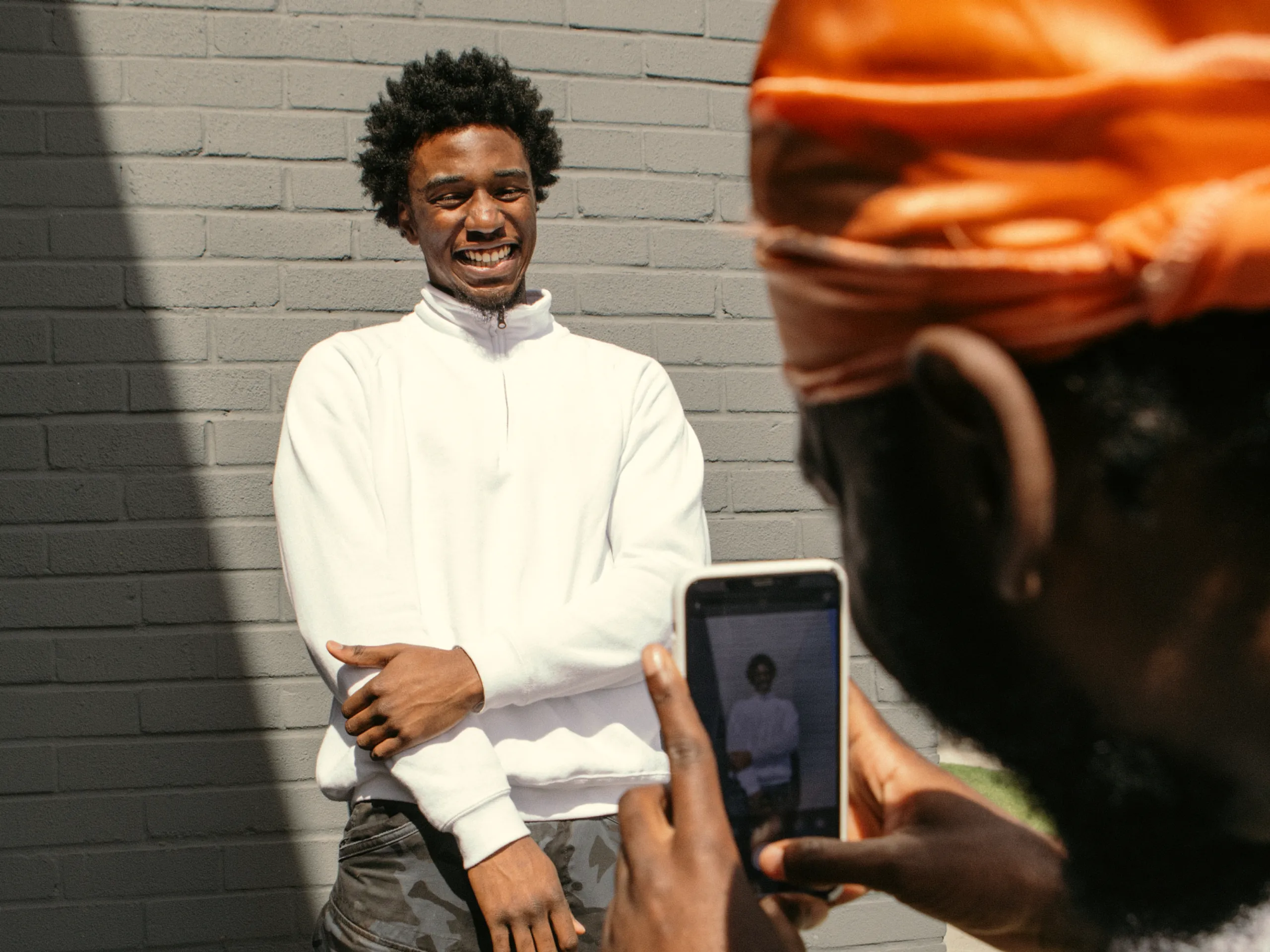 A person in a white sweatshirt smiles while standing against a gray brick wall as another person takes their photo with a smartphone.