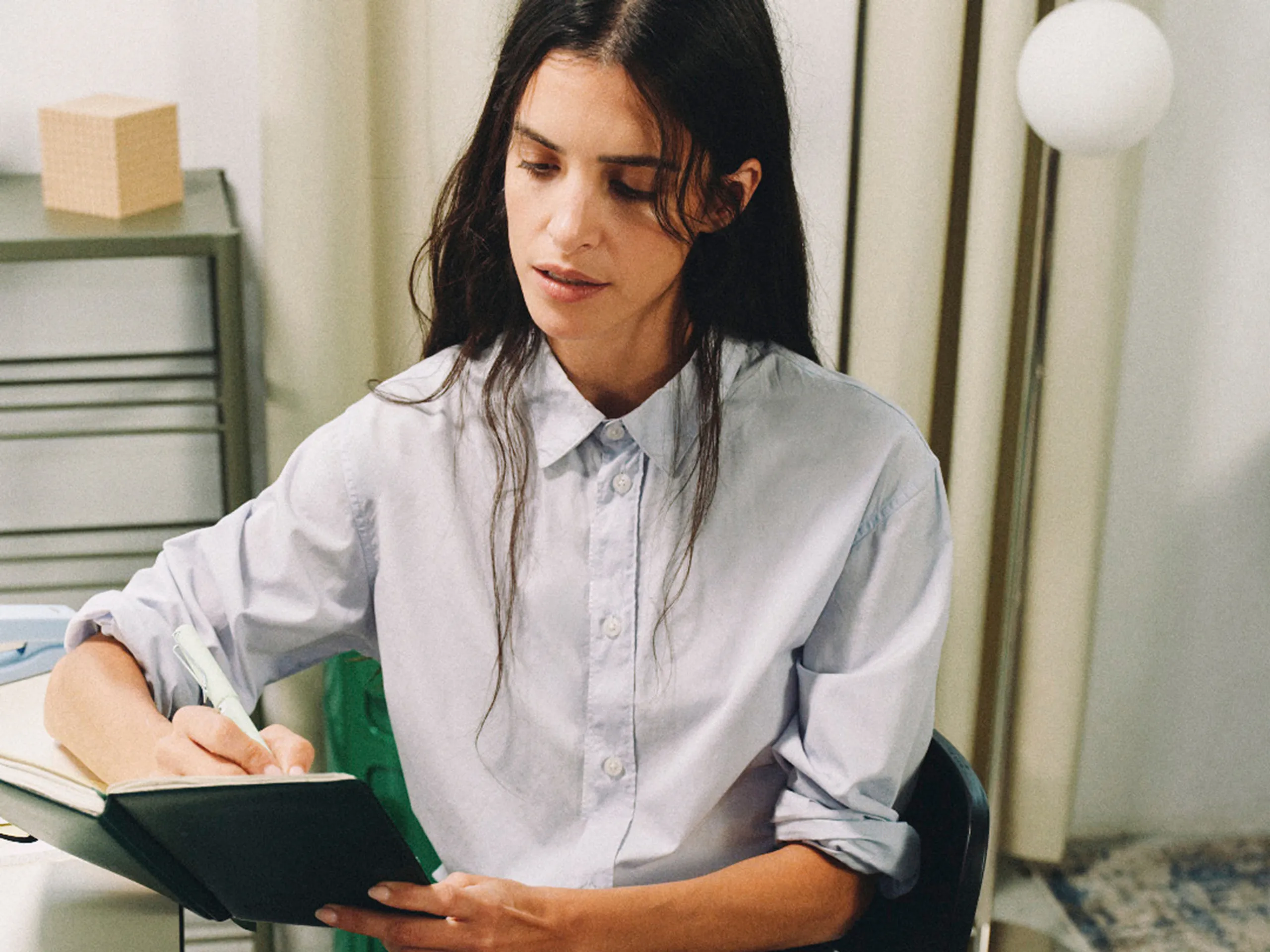 A woman with long, dark hair wearing a light blue button-down shirt sits at a desk, focused on writing in a dark-covered notebook with a light-colored pen. The setting appears to be a brightly lit, modern office or workspace.