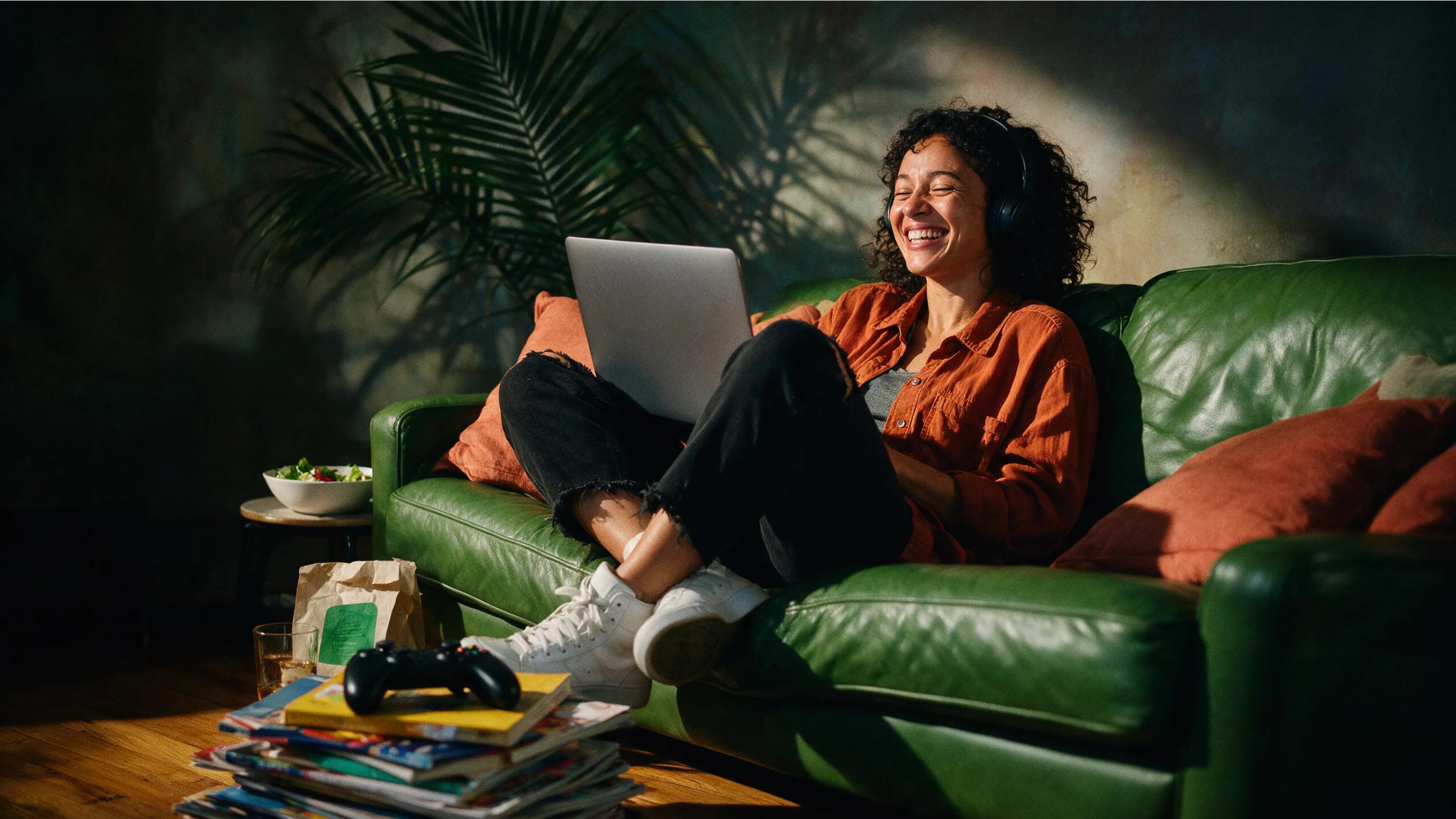 A woman wearing headphones laughs while using a laptop on a green couch.
