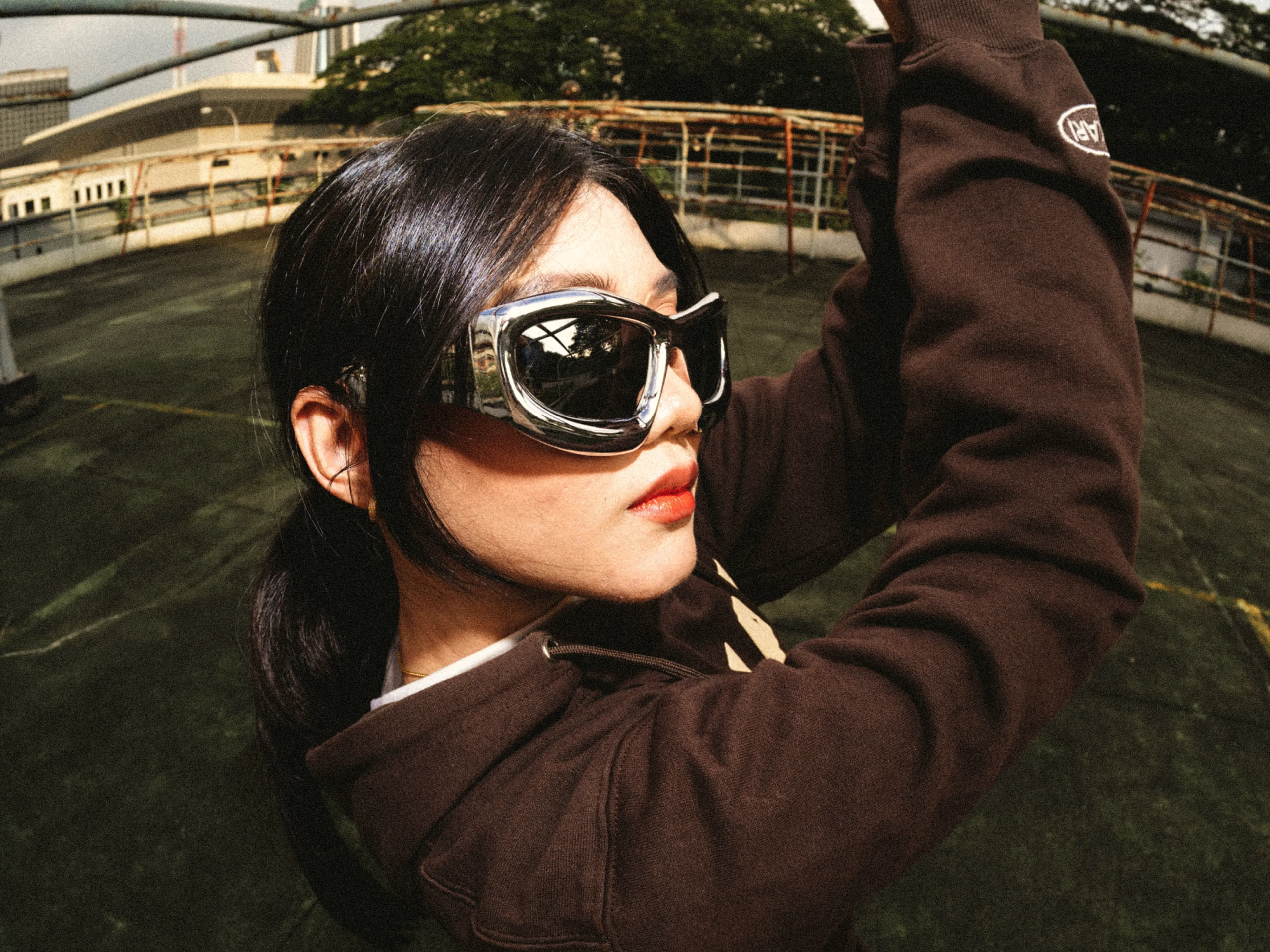 A person with dark hair and red lipstick wears large, reflective sunglasses and a brown jacket, posing outdoors on a rooftop with green trees and buildings in the background.