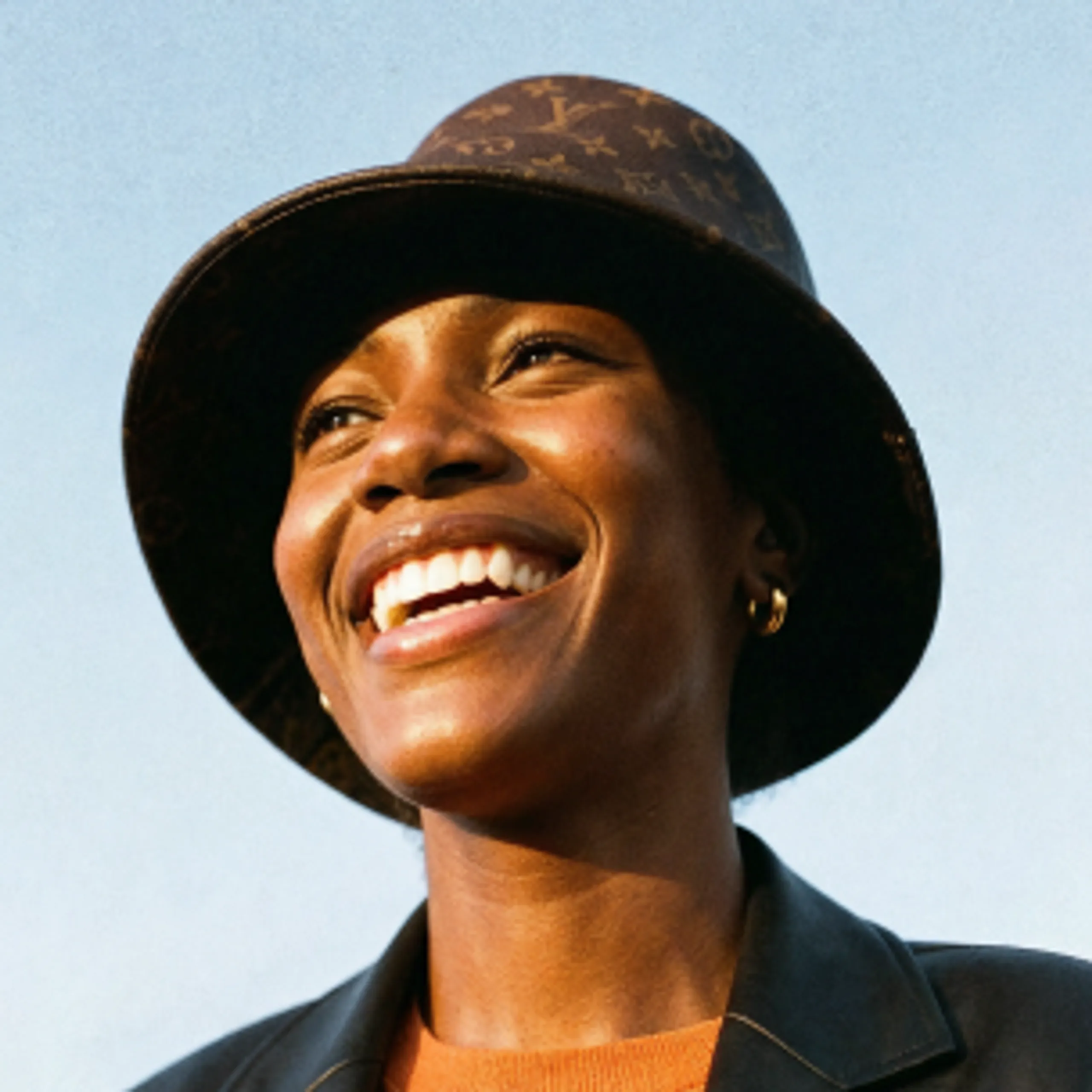 A person smiling widely, wearing a dark bucket hat with a patterned design, a dark jacket, and an orange top, standing against a clear blue sky.