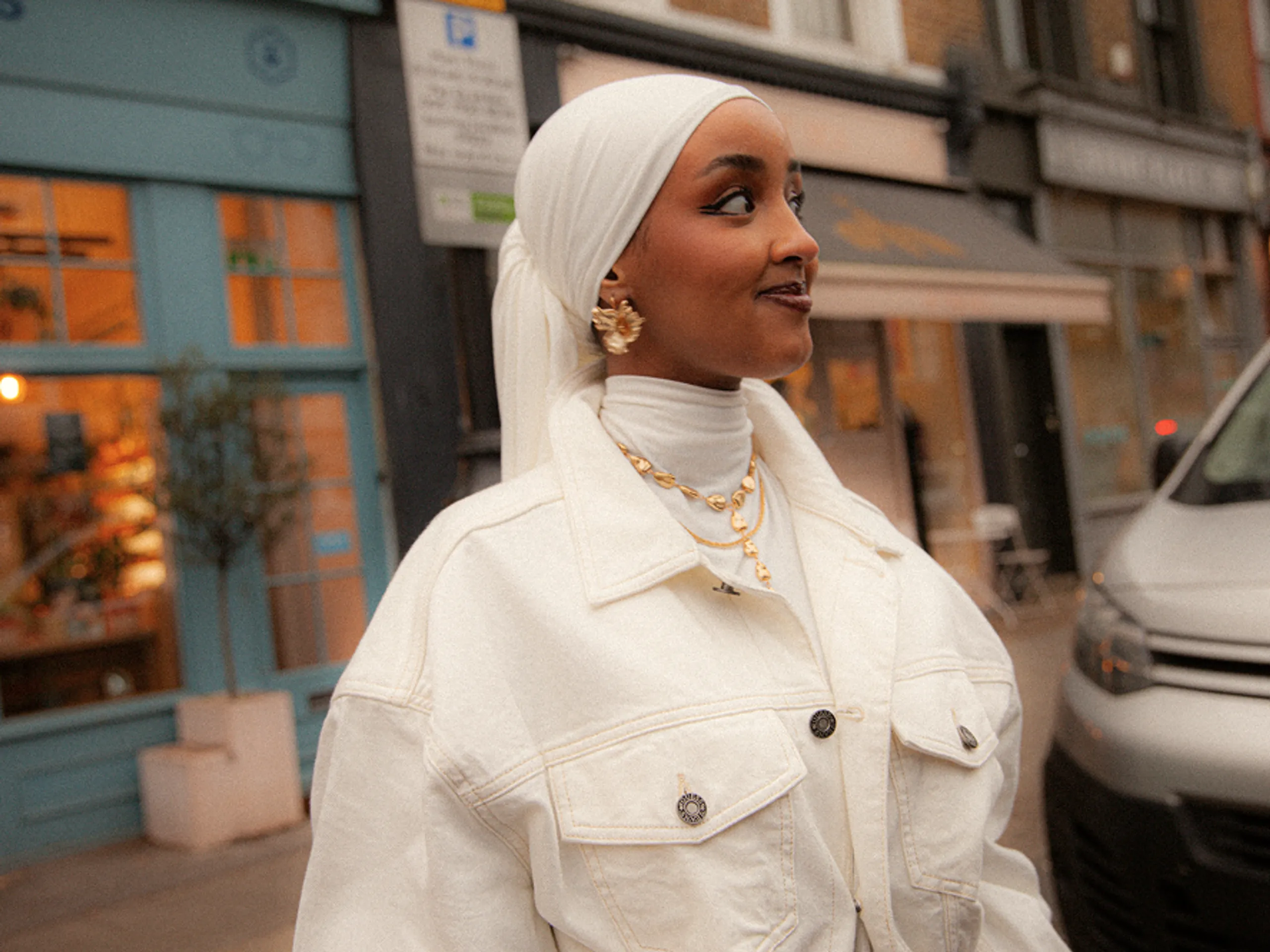 A woman in a white hijab and white jacket smiles while standing on a city street, with buildings and a parked car in the background.