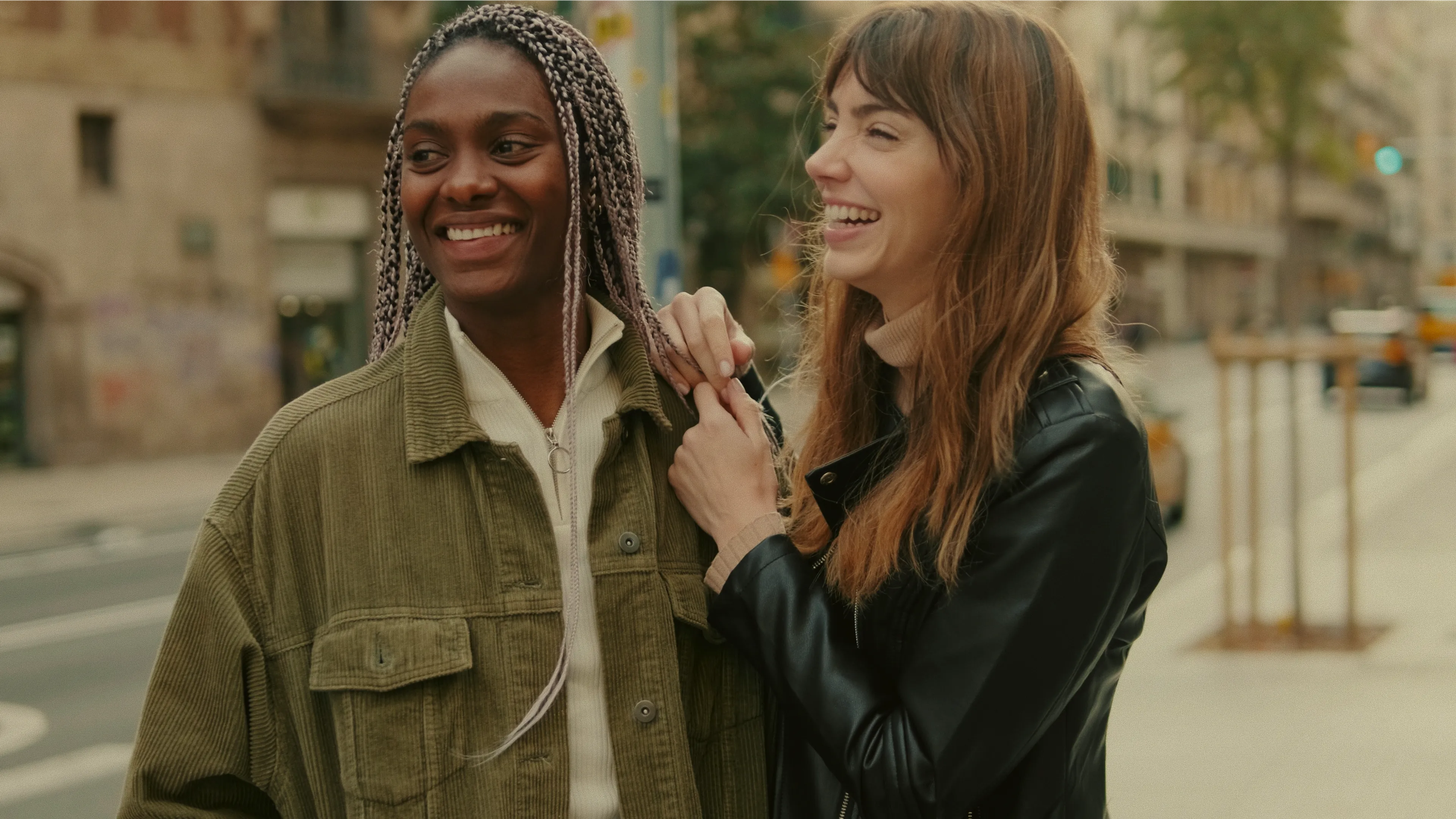 Two young women stand outdoors on a city sidewalk, smiling and laughing together. One has braided hair and wears an olive jacket; the other has long brown hair and wears a black jacket.