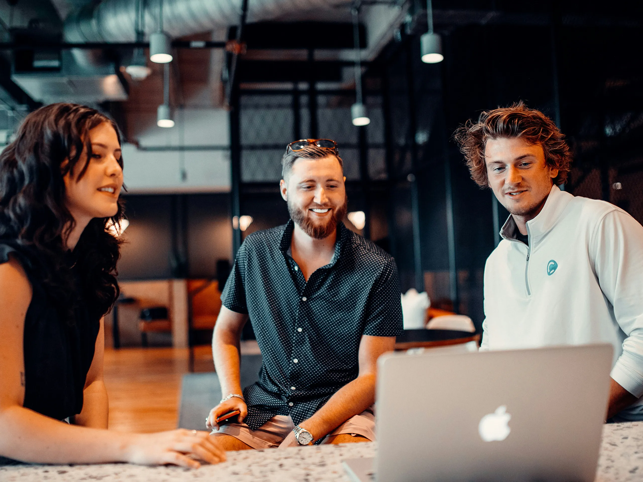 Three young adults sit at a table, smiling and looking at a laptop screen in a modern, industrial-style office space.