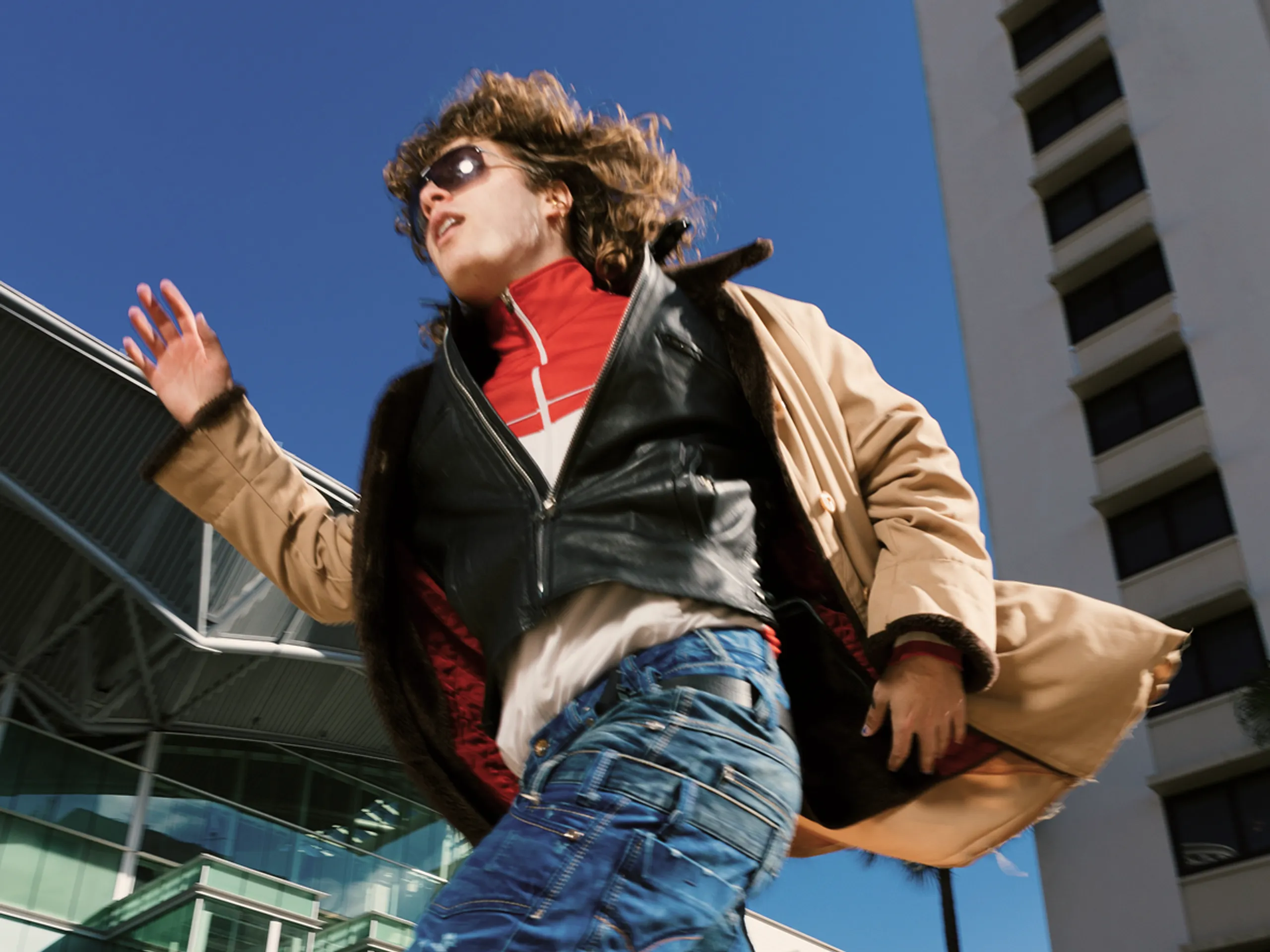 A person wearing sunglasses, a tan coat, and blue jeans runs outdoors by modern buildings under a clear blue sky.
