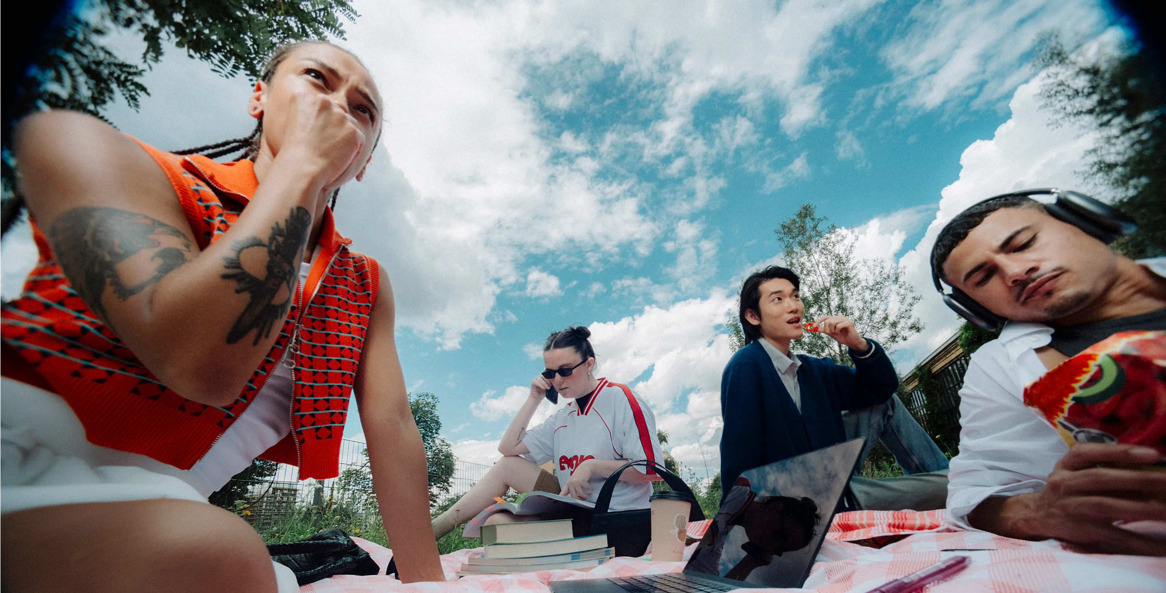 Four people relax on a picnic blanket outdoors. One eats chips, another listens to headphones, one holds a phone, and another appears to talk. Laptops and books are scattered nearby under a blue sky with clouds.