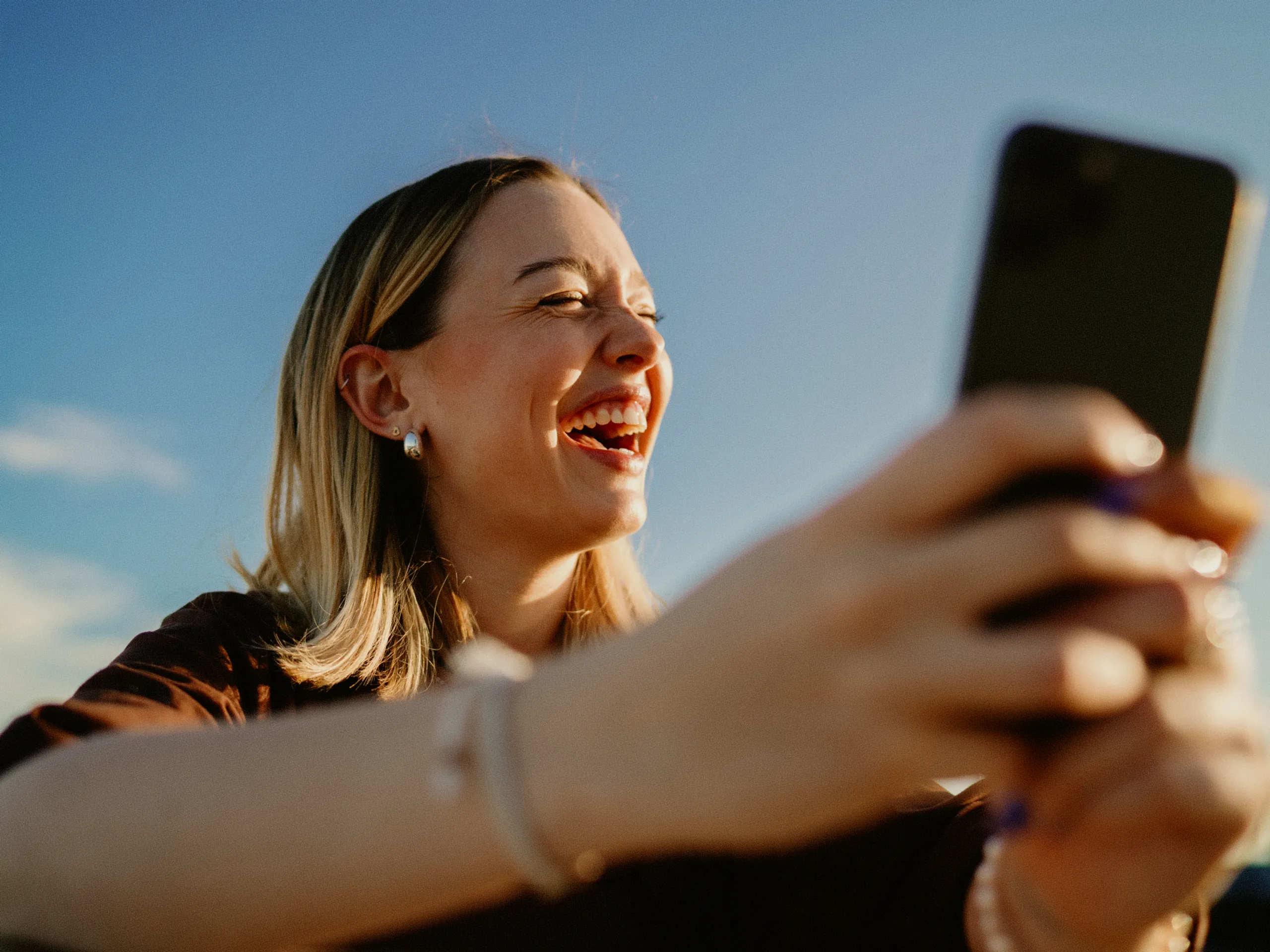 A young woman smiles and laughs while taking a selfie outdoors, holding a phone in her outstretched hand against a clear blue sky.