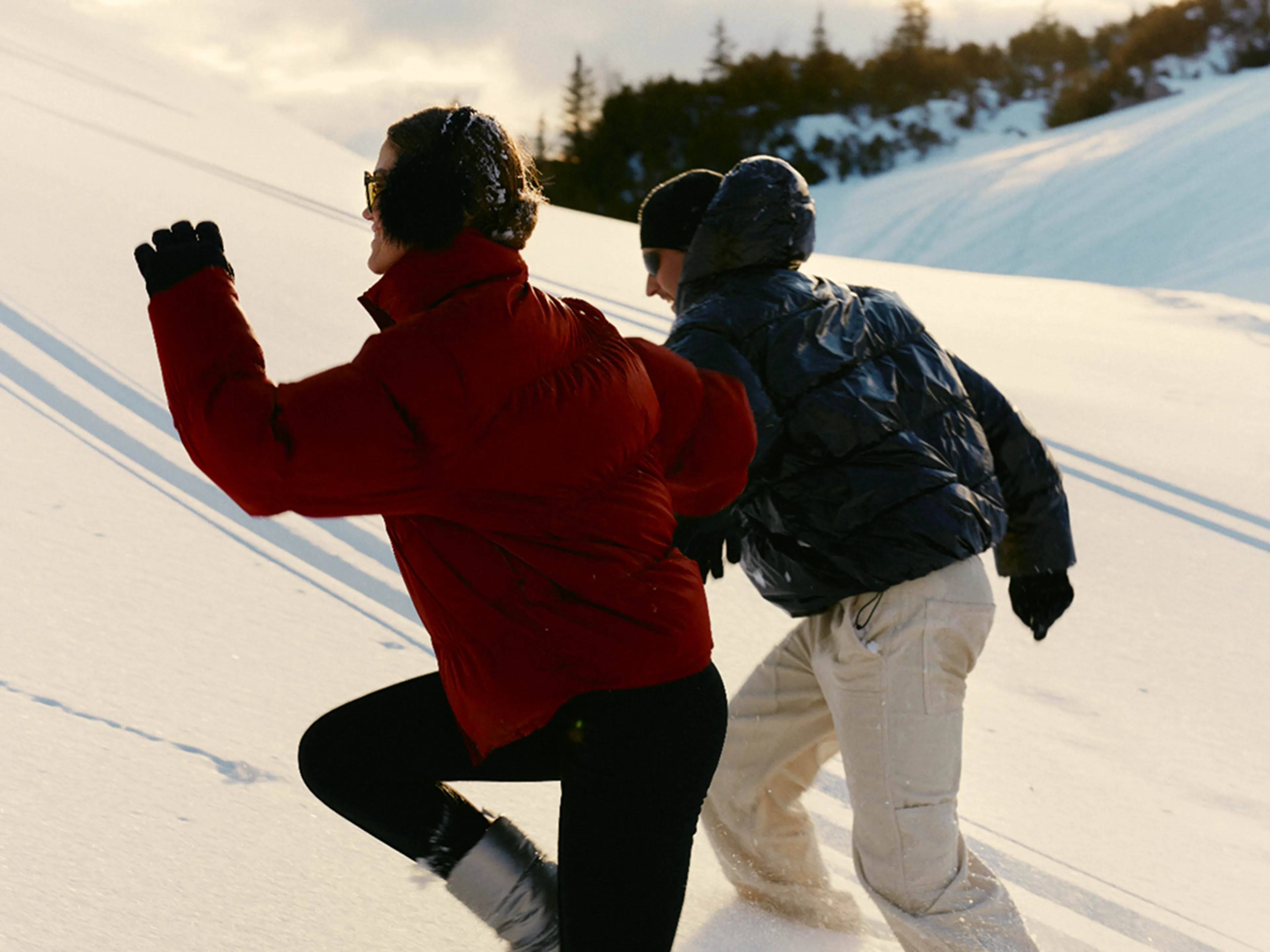 Two people dressed in winter clothing run up a snowy hill, leaving footprints behind them. Pine trees and a cloudy sky are visible in the background.