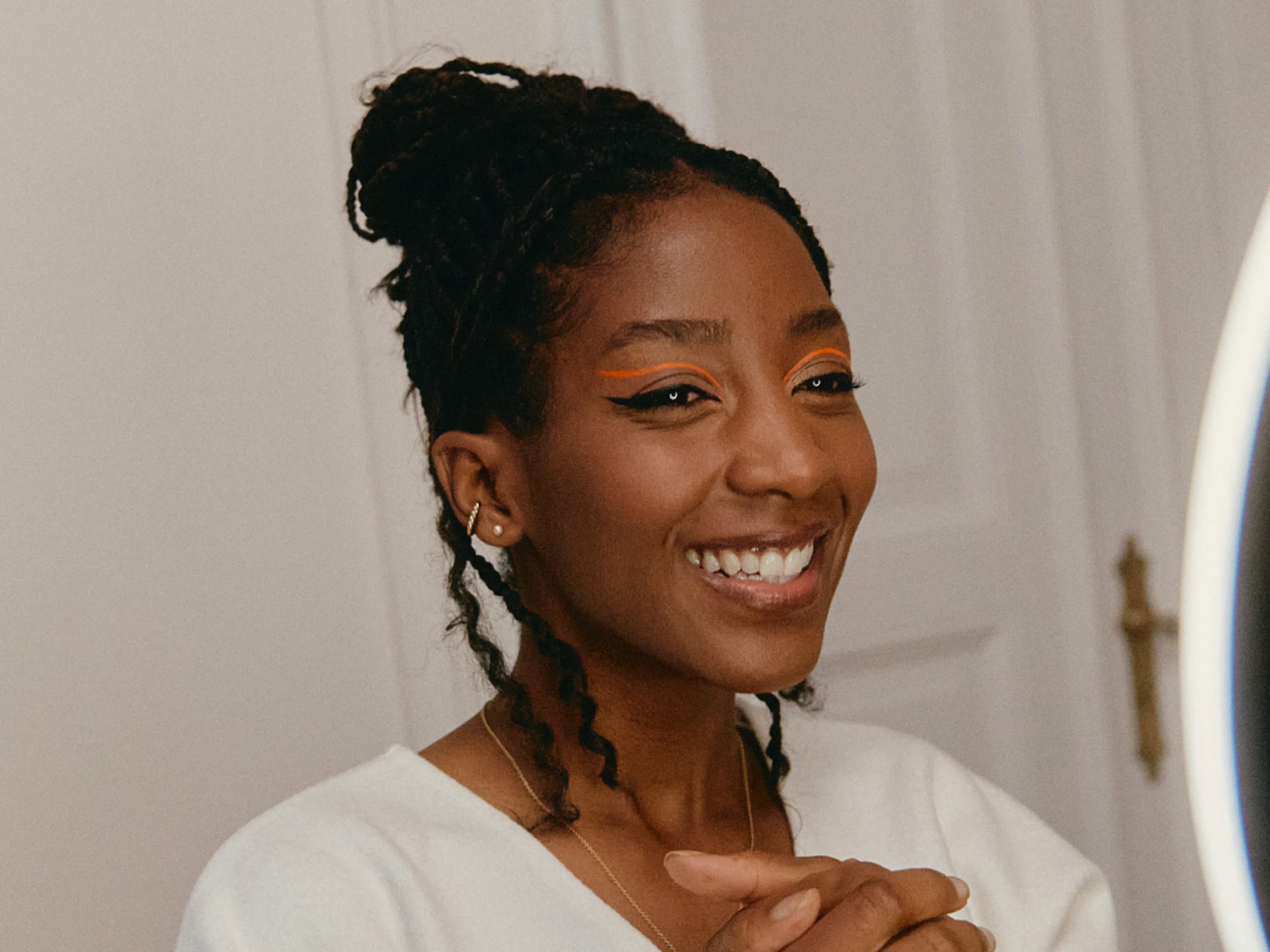 A woman with braided hair smiles in front of a mirror. She wears bold orange and blue eyeliner and a white top, standing indoors with a door and wall in the background.