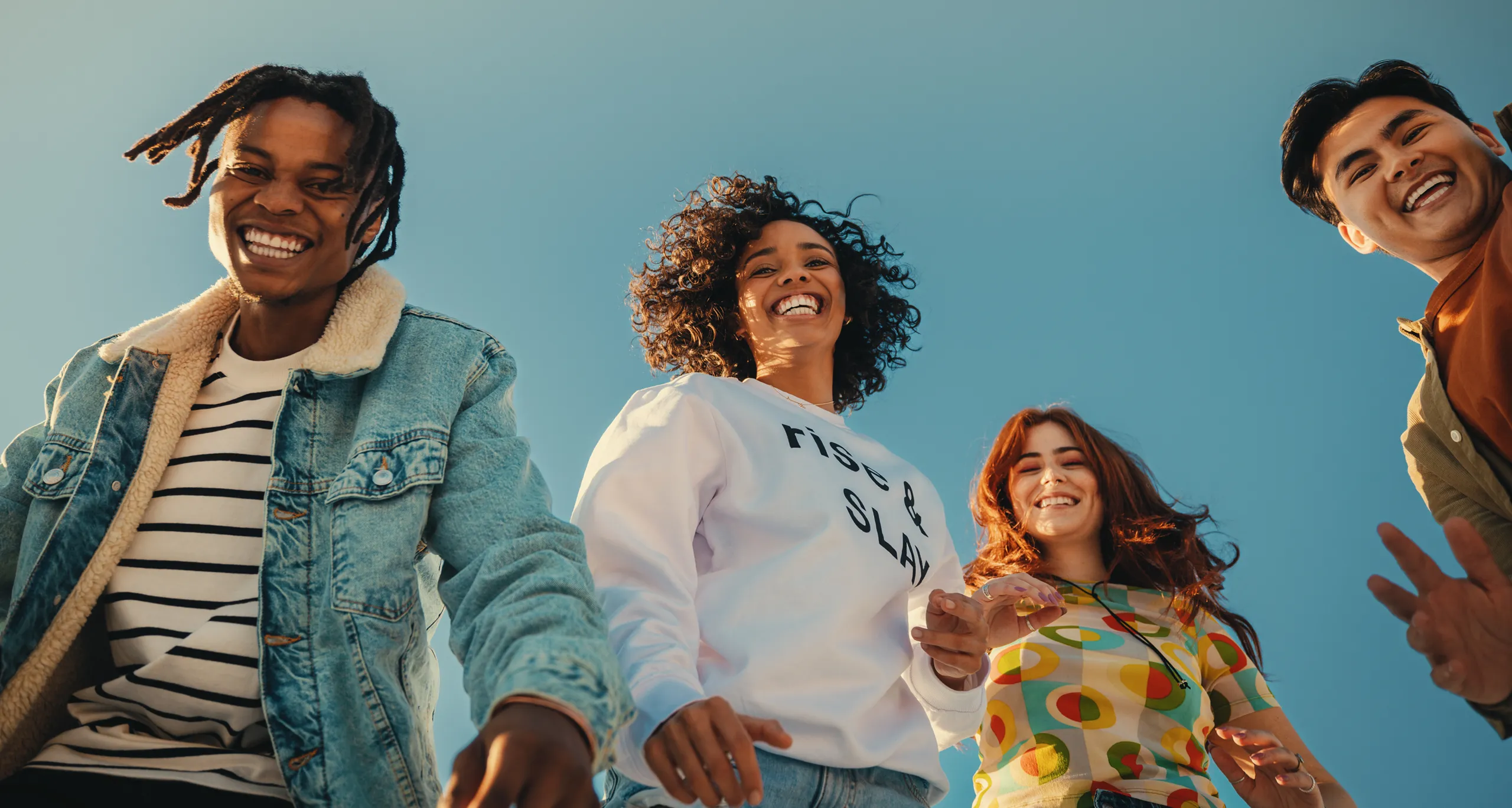 Four young adults smile and laugh together outdoors, standing close and looking down toward the camera against a clear blue sky.