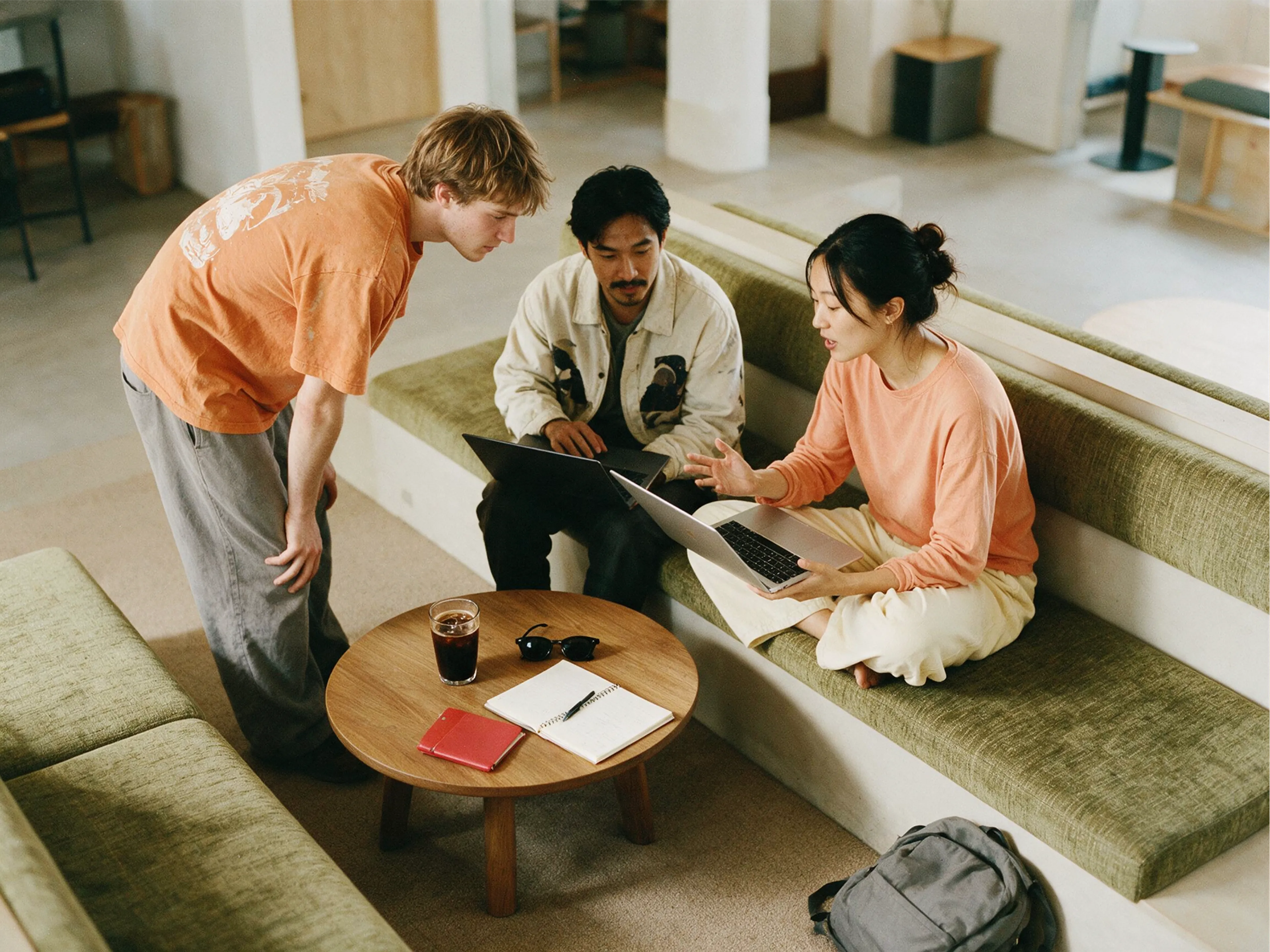 Three people are gathered around a coffee table with laptops, notebooks, and a drink, collaborating in a modern, casual workspace with green couches and soft lighting.