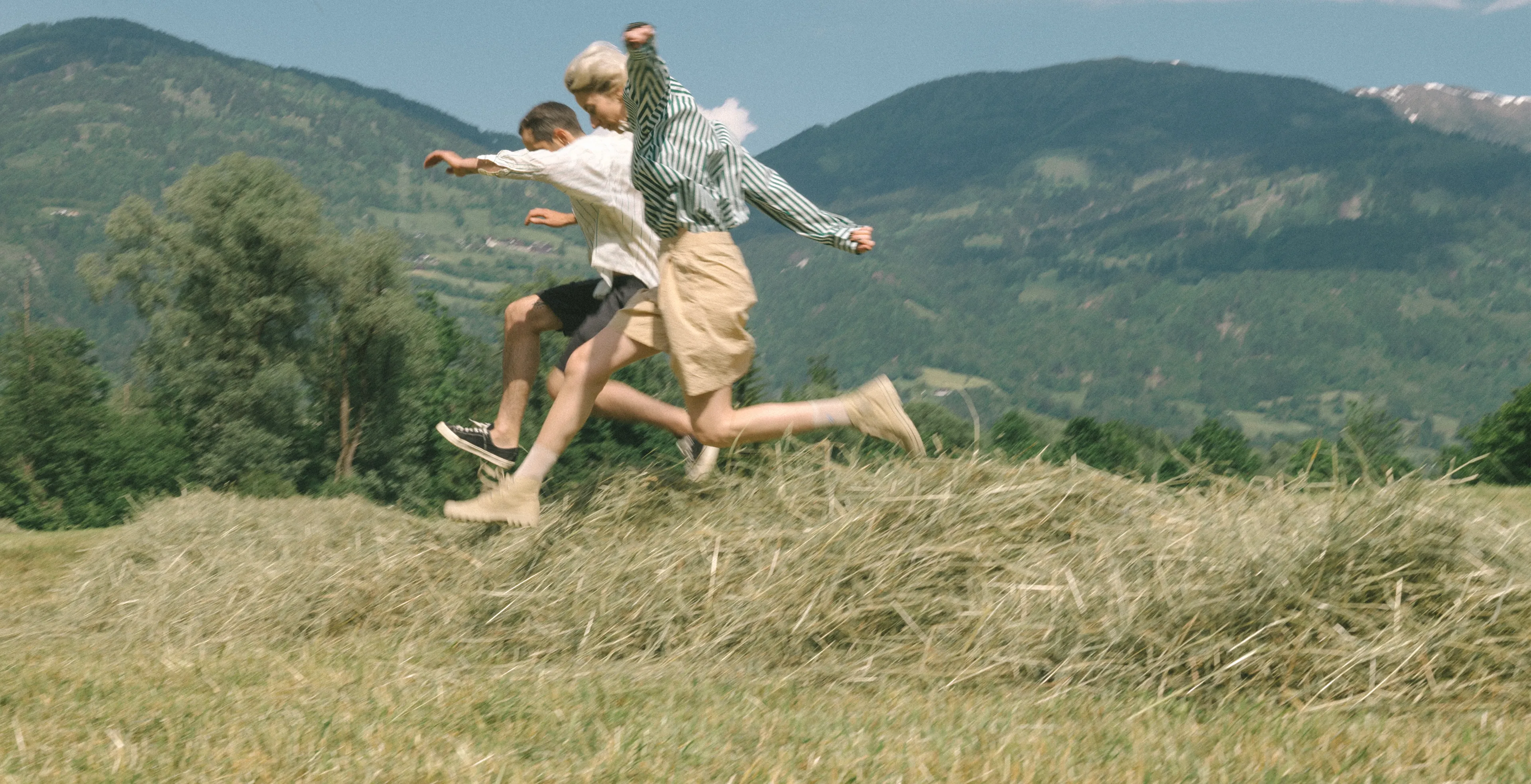 Two people are jumping over a pile of hay in a grassy field, with green trees and distant mountains in the background under a clear blue sky.