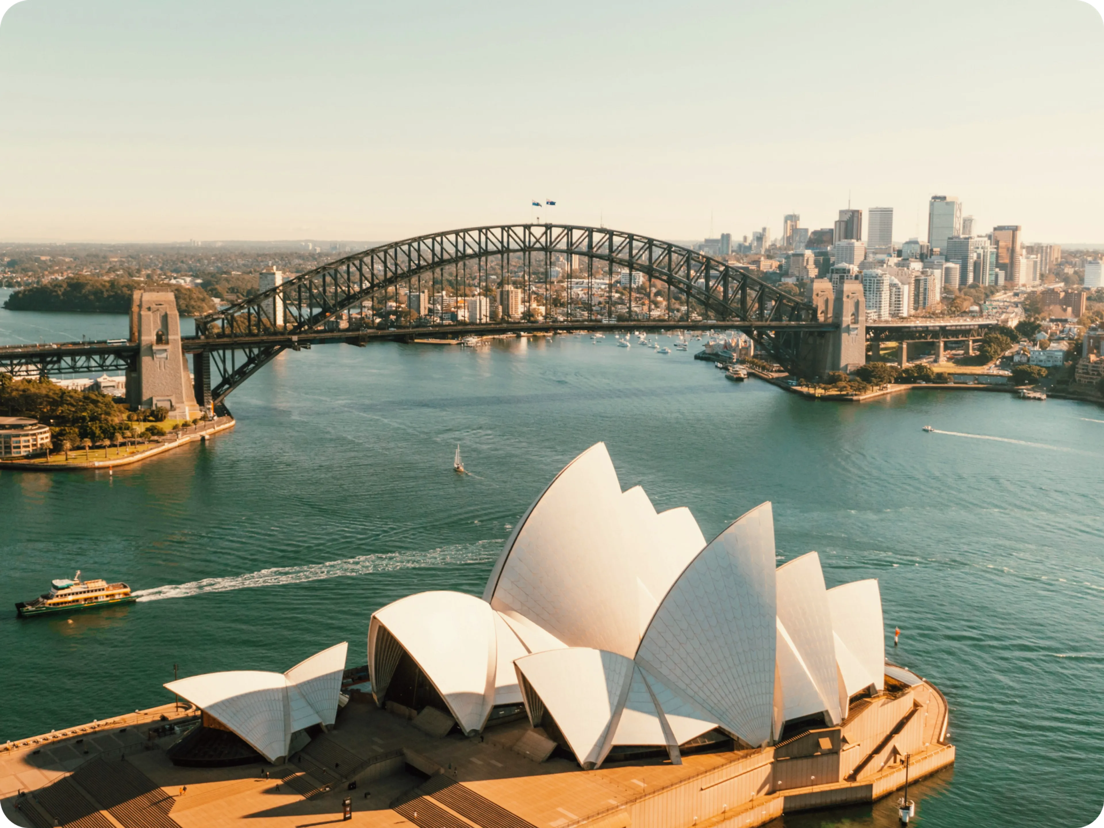 Aerial view of the Sydney Opera House in the foreground with the Sydney Harbour Bridge and city skyline in the background on a sunny day.