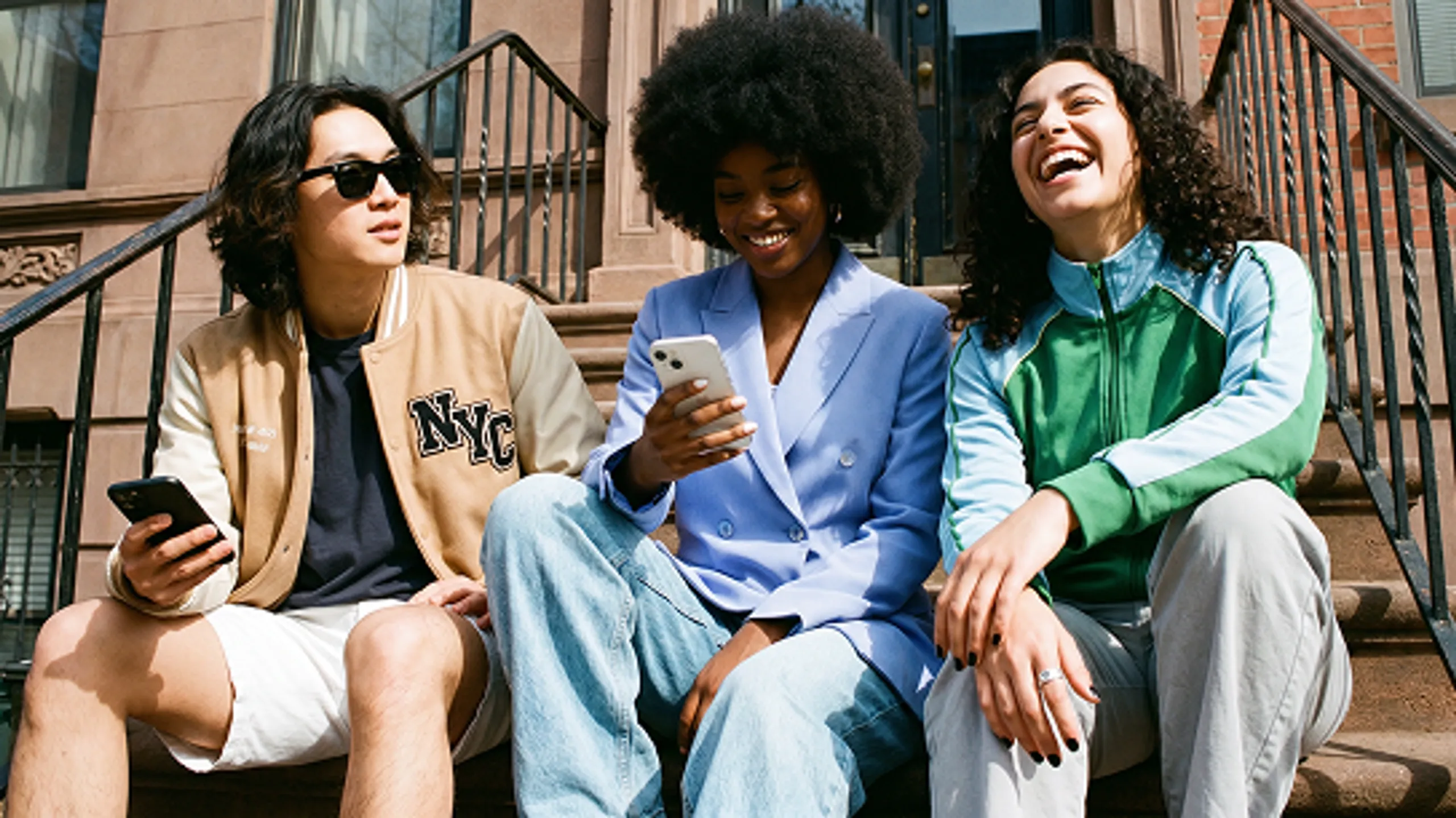 Three friends sit on outdoor steps, smiling and laughing. Two hold smartphones, one in a tan NYC jacket, one in a blue blazer, and the third in a green jacket. The background shows a brownstone building.