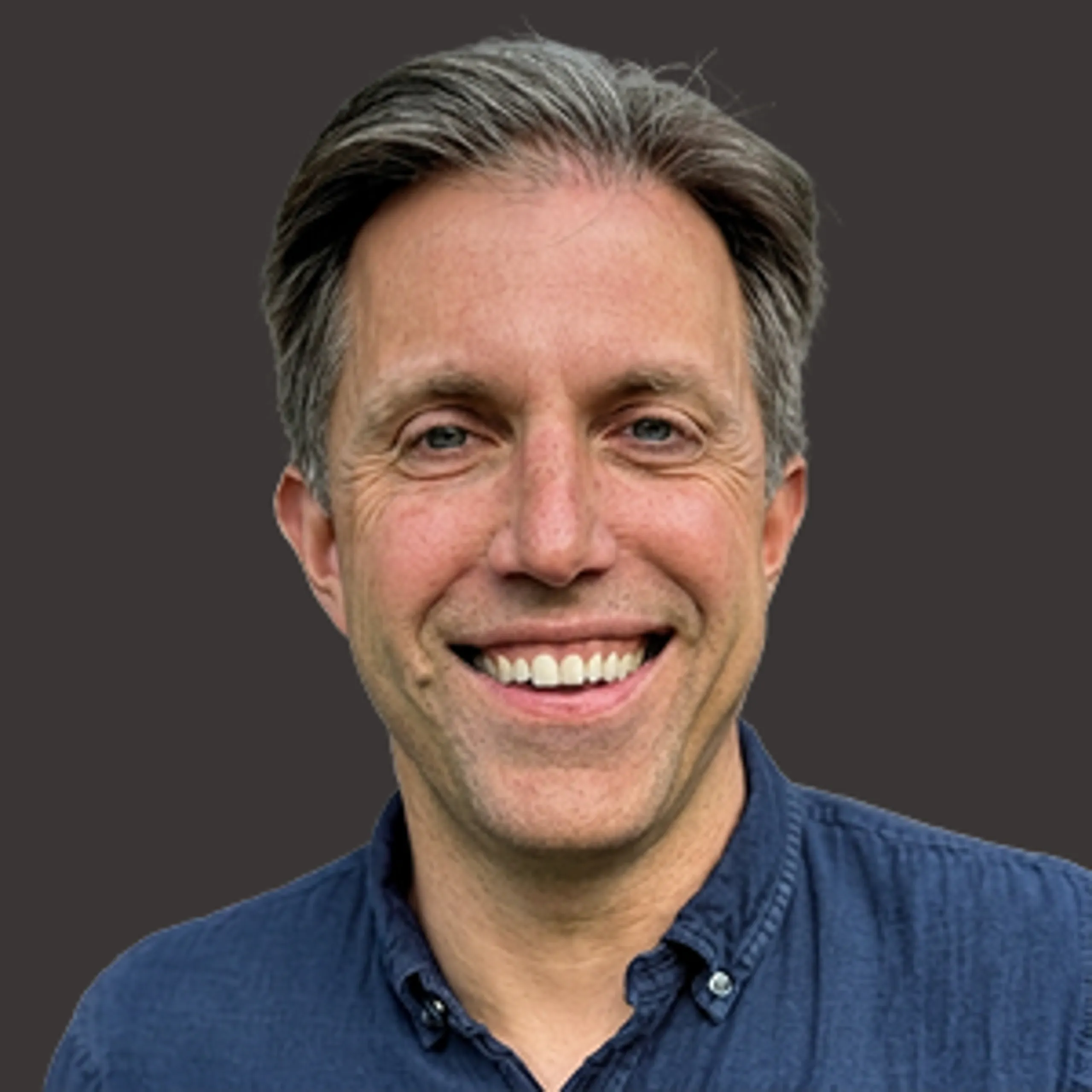 Smiling man with short grayish-brown hair wearing a dark blue button-up shirt, standing against a plain dark background.