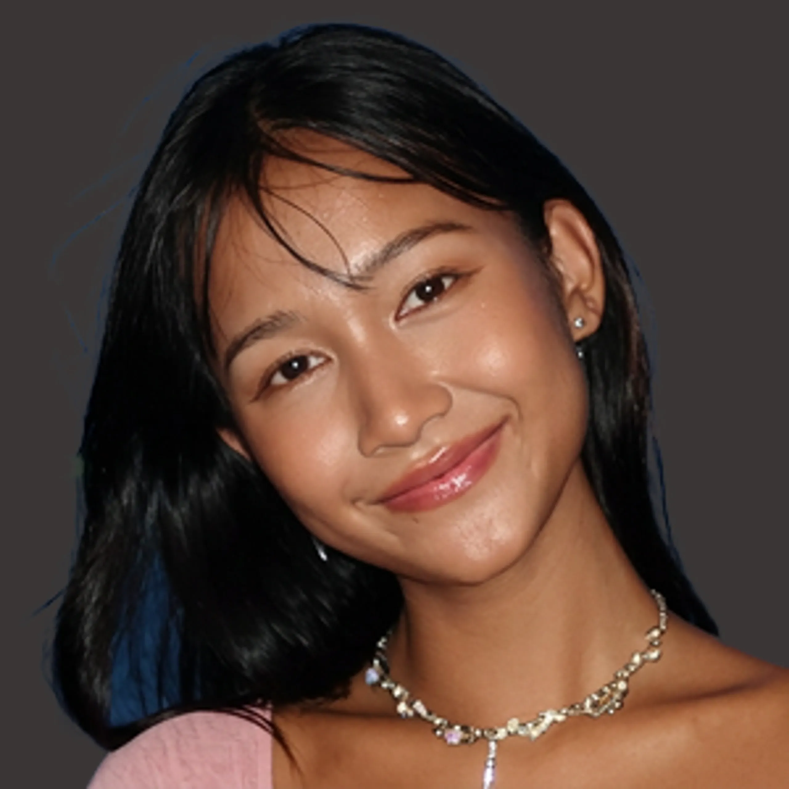 A young woman with straight black hair smiles softly, wearing a pink top and a silver necklace, in front of a dark background.