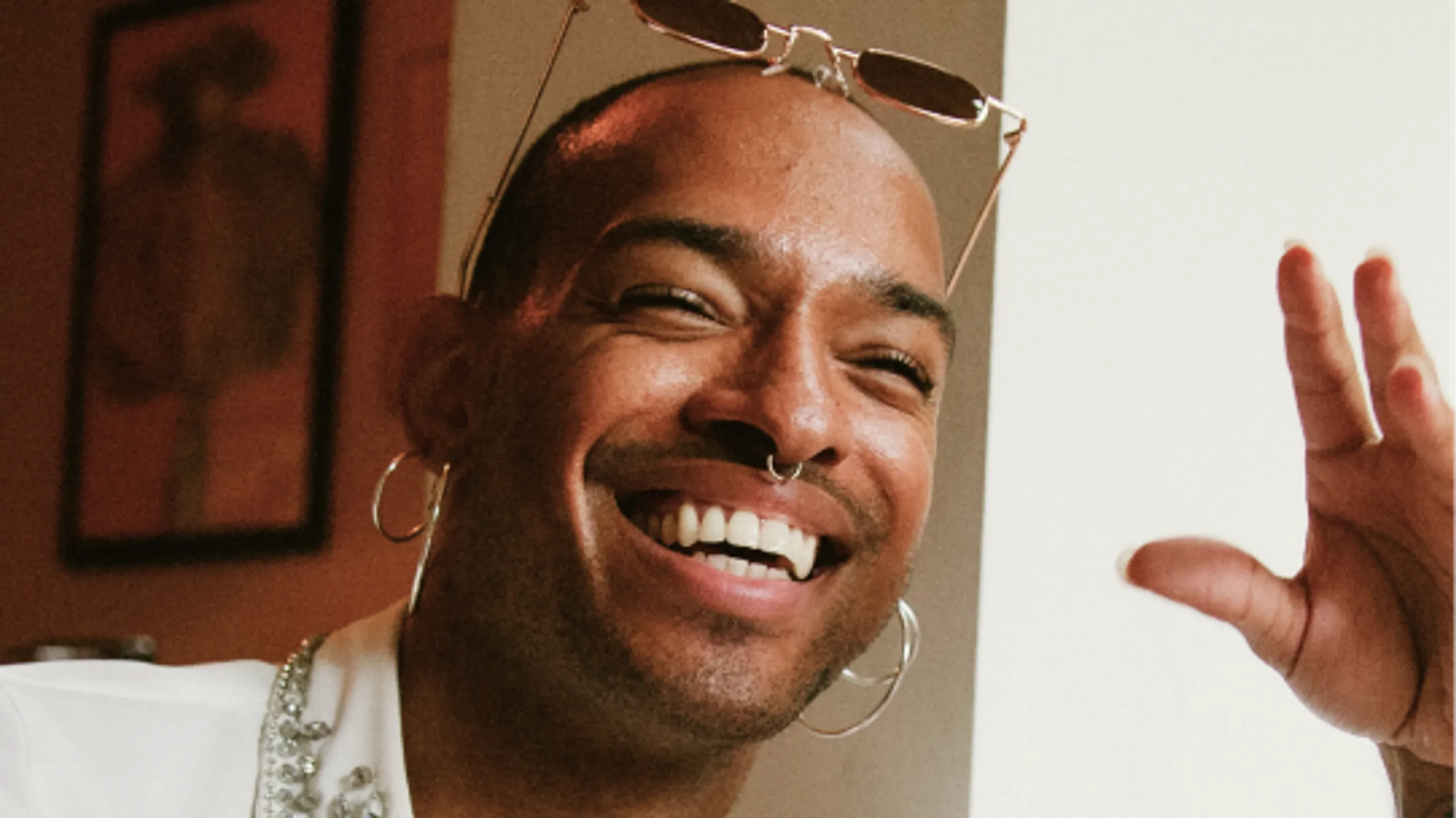 A smiling person with glasses on their head, hoop earrings, and a nose ring, raising their hand in a friendly gesture. The background is softly lit with a framed picture on the wall.