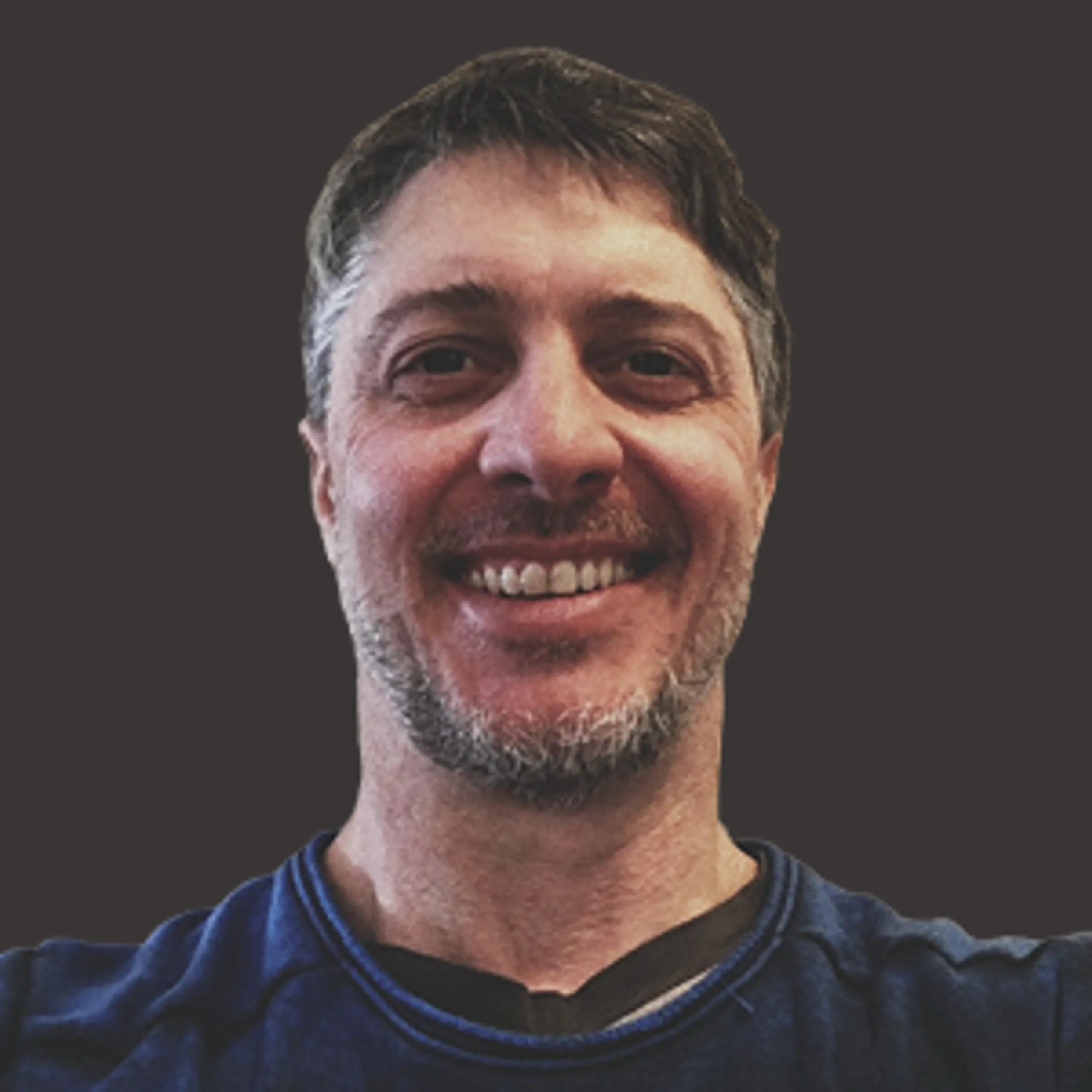 Smiling man with short dark hair and a gray beard, wearing a blue shirt, in front of a plain dark background.