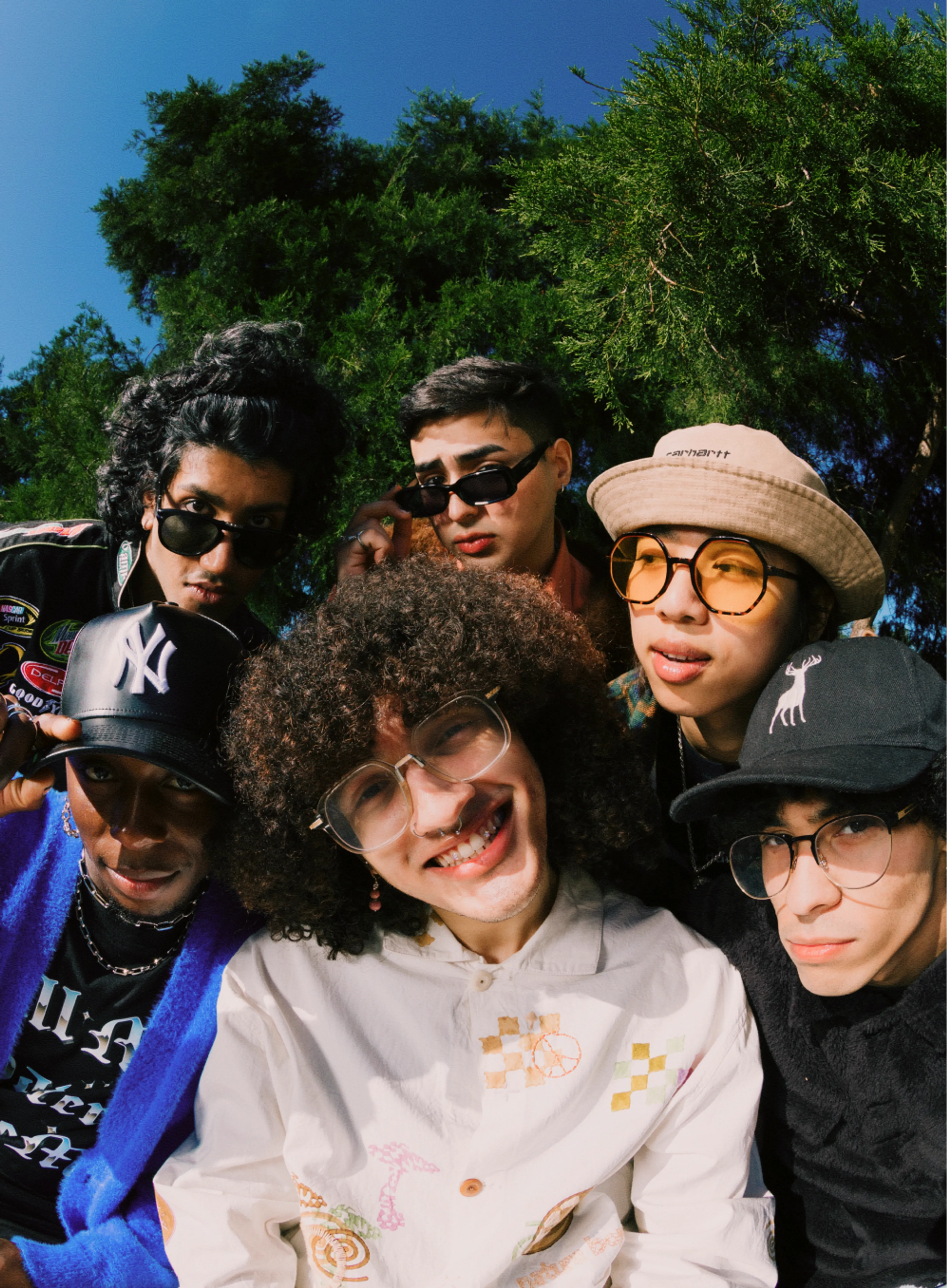 Six young people, dressed in trendy clothes and sunglasses, pose closely together outdoors in front of green trees under a clear blue sky, smiling and looking at the camera.