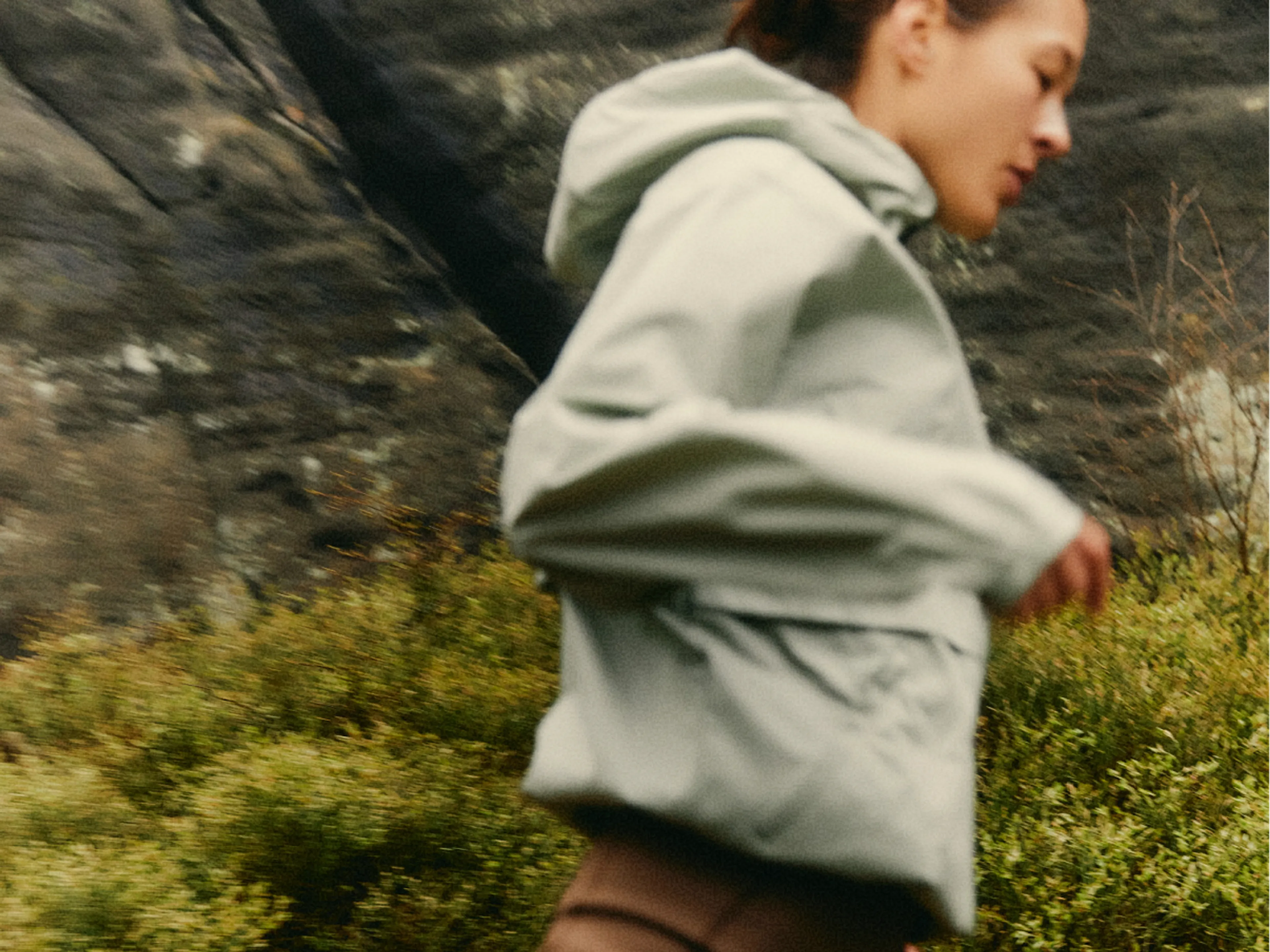 A person in a light jacket and brown pants moves outdoors, with blurred motion suggesting activity. Green shrubs and a rocky background are visible.