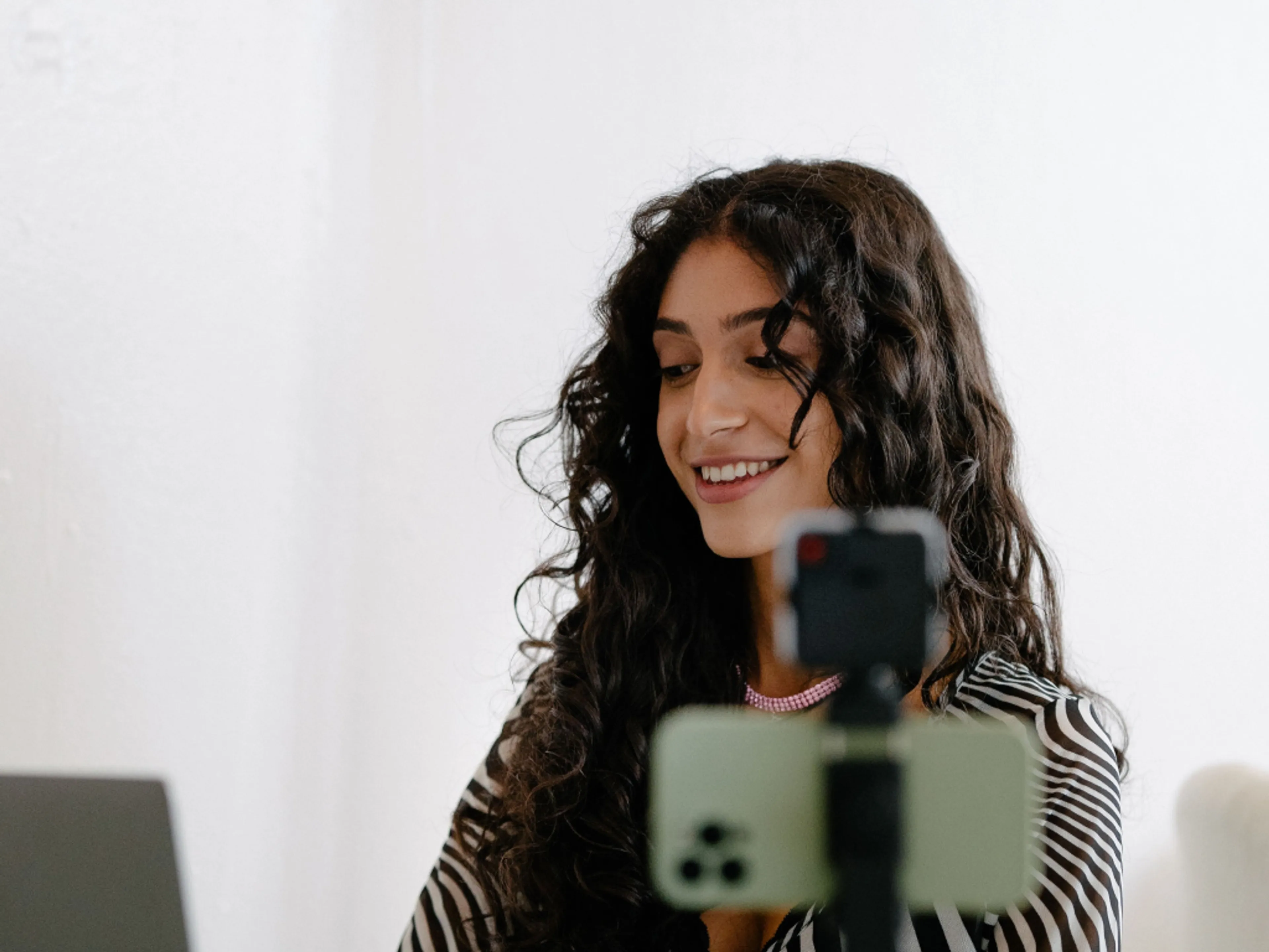 A woman with long curly hair smiles while looking at a laptop. A smartphone on a tripod is set up in front of her, suggesting she is recording or live streaming in a bright, indoor setting.