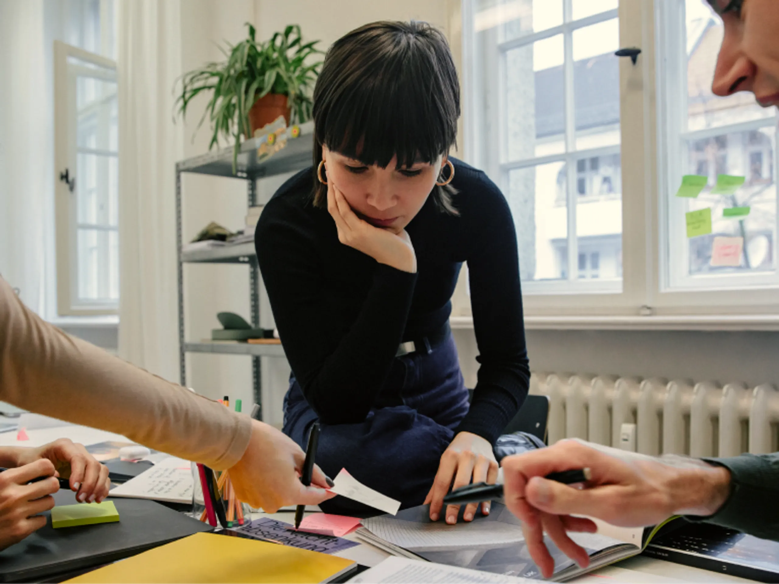 A woman leans over a desk, looking at papers while two other people point at documents and notes. The scene appears to be a collaborative work or study session in a bright office space.