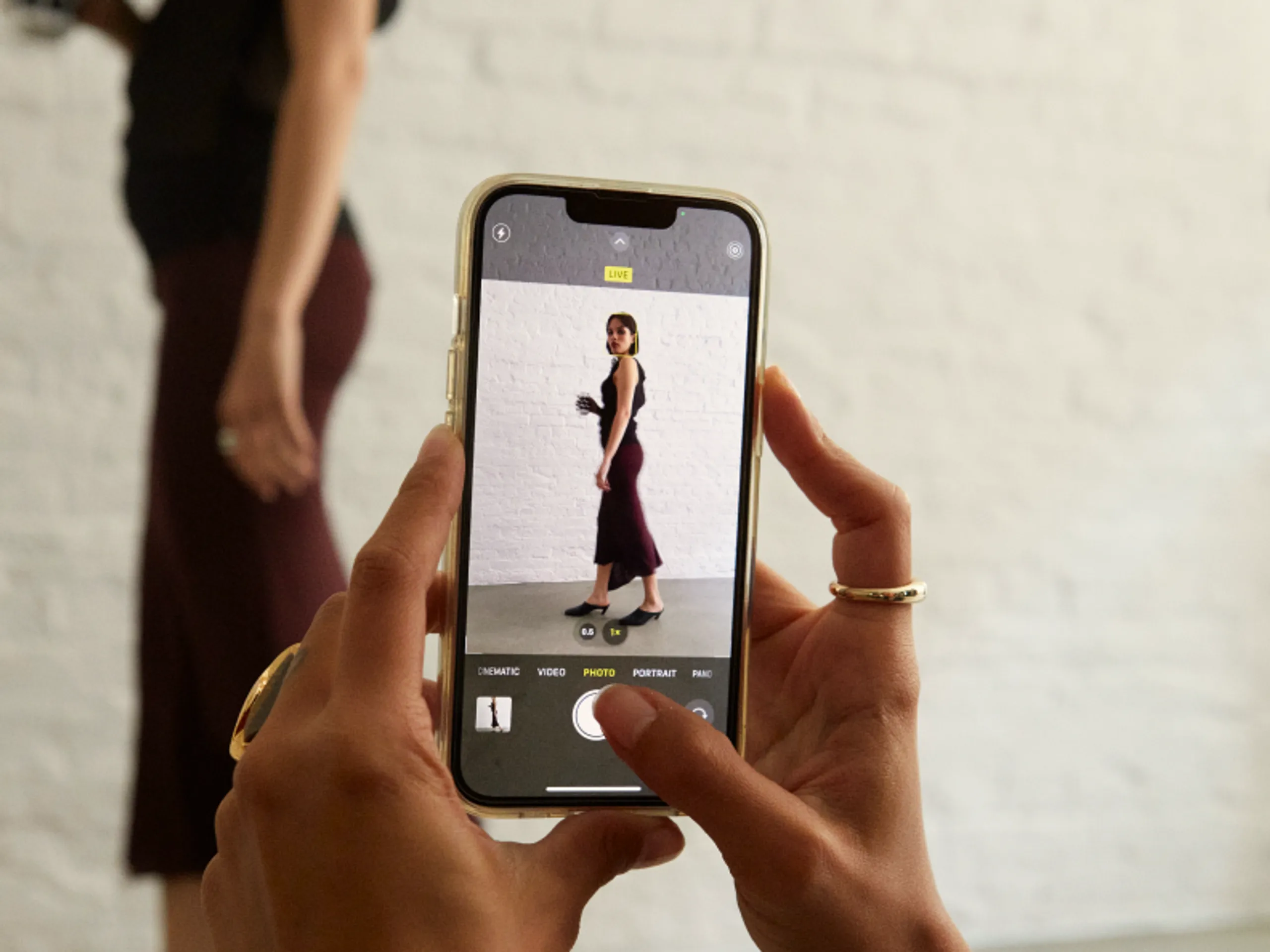 A person holds a phone, taking a photo of a woman in a sleeveless top and skirt standing against a white brick wall. The camera app is open, focusing on the woman.